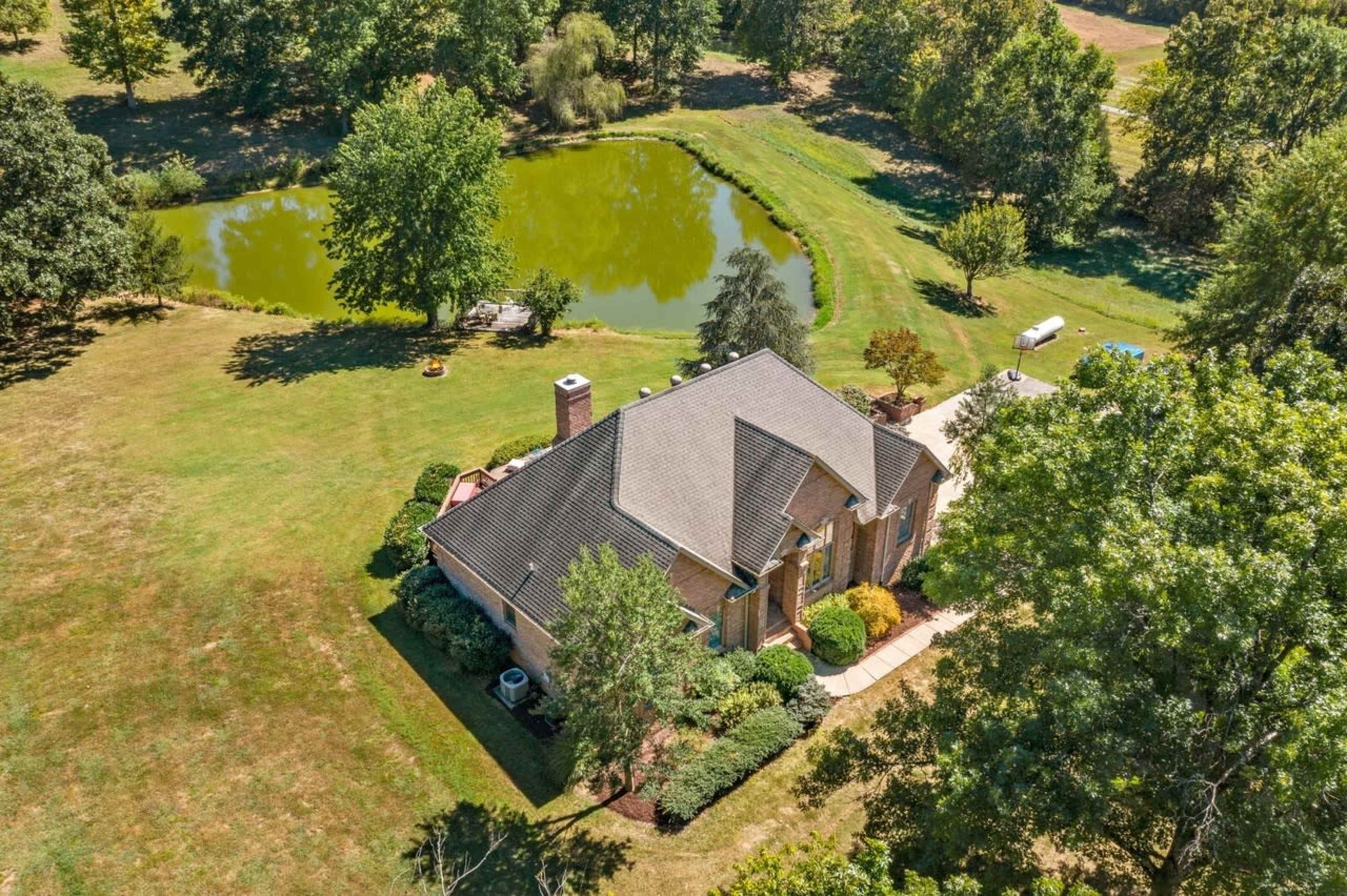 The image shows a single-story brick house nestled among trees, overlooking a small pond in a green, open landscape.