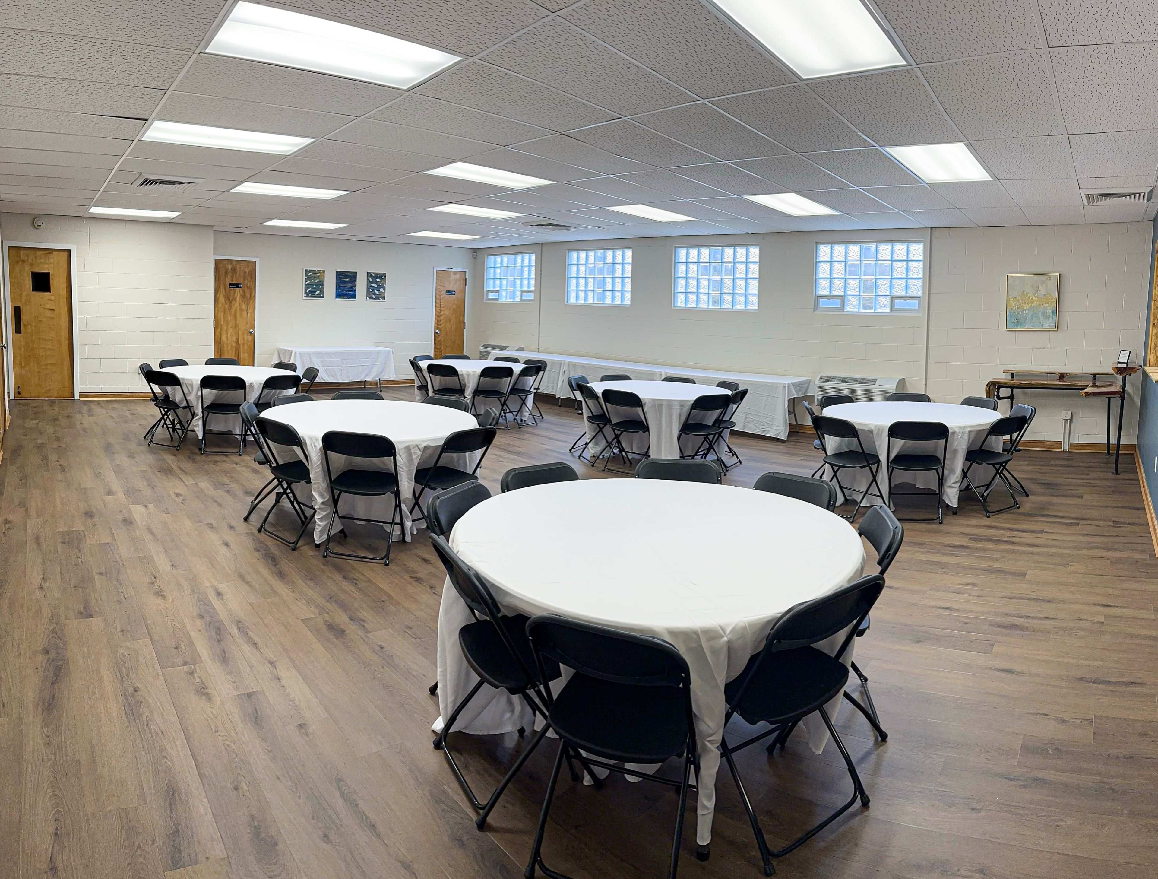 A spacious room set up for an event, featuring multiple round tables covered with white tablecloths and surrounded by black chairs.