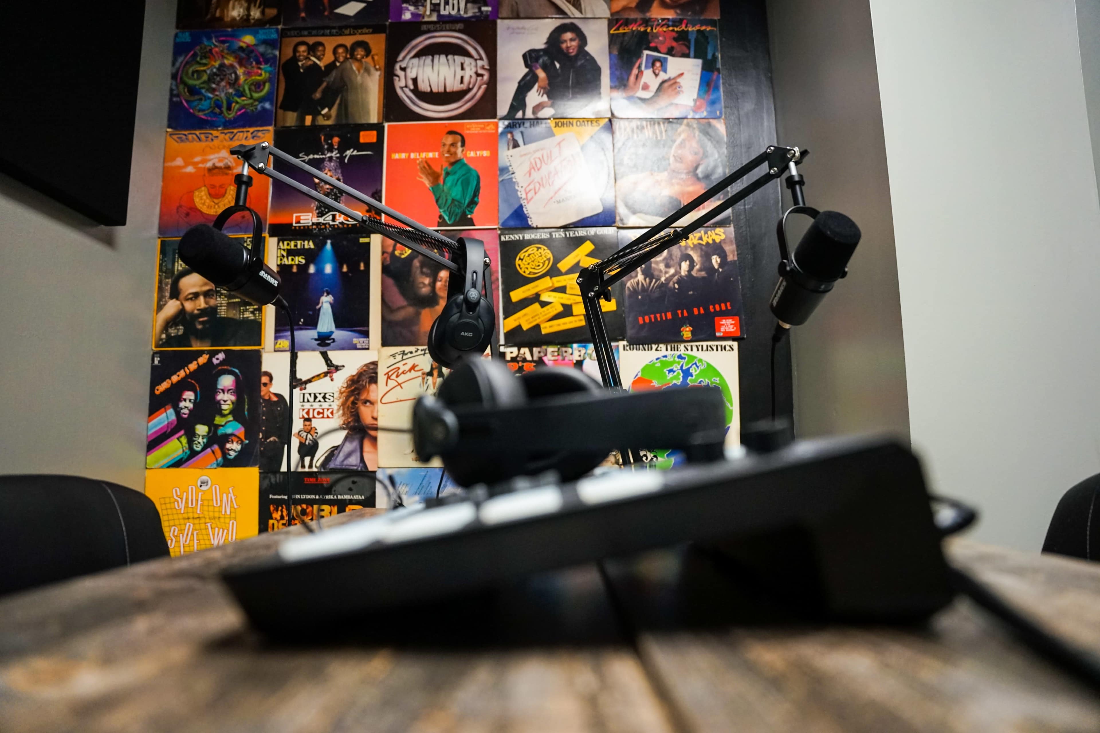 The image shows a podcast recording setup featuring two microphones on adjustable arms in front of a wooden table, with a wall decorated in colorful album covers.