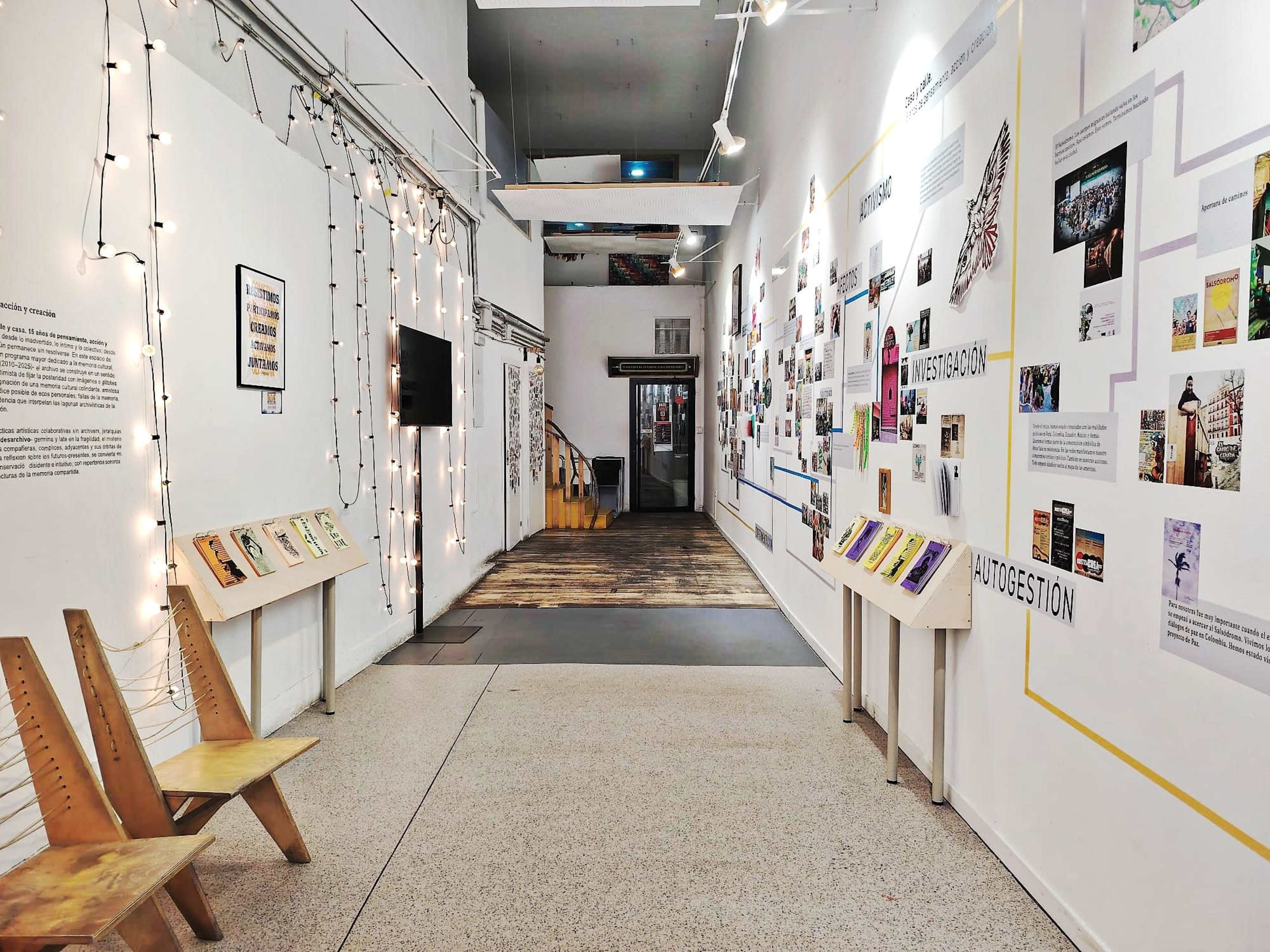 The image shows a hallway in a gallery with walls decorated with various artworks, information panels, and illuminated by string lights.