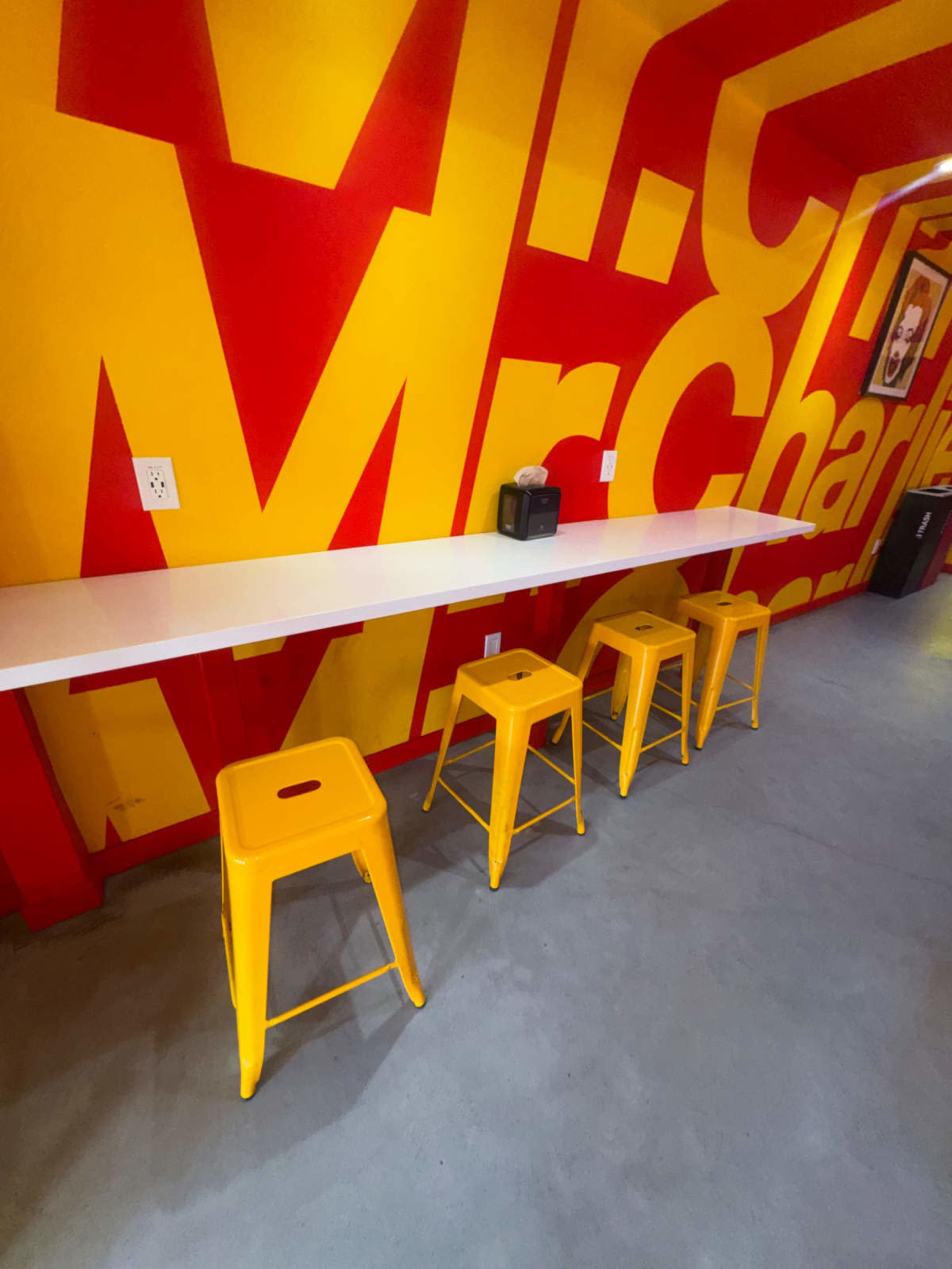 A narrow, white countertop with four yellow stools is set against a vibrant red and yellow wall featuring the repeated name "Mr. Charlie."