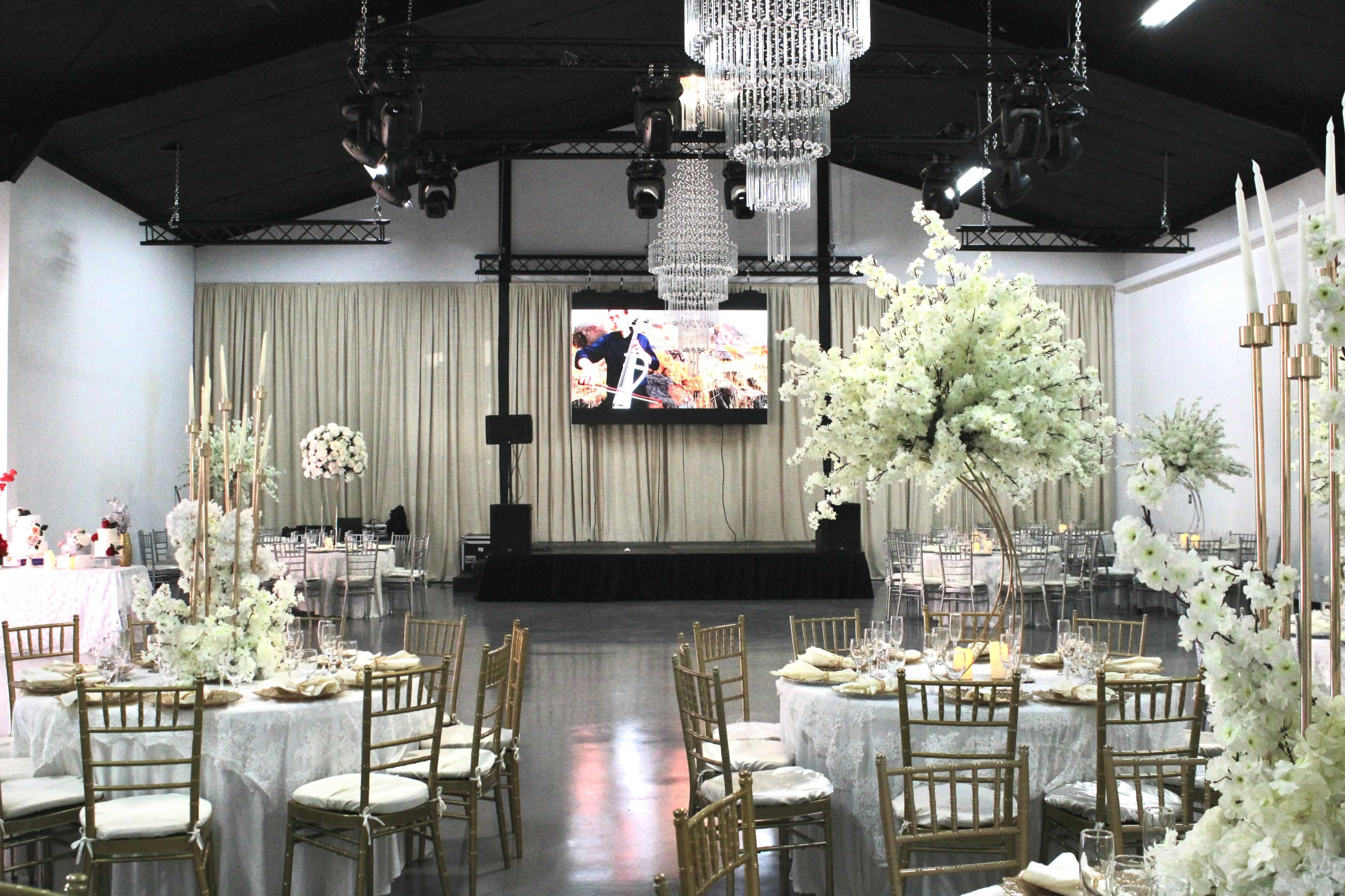 An elegantly arranged event space with round tables set for a gathering, decorated with white floral centerpieces and gold chairs, and features a large screen displaying visuals on a staged platform.