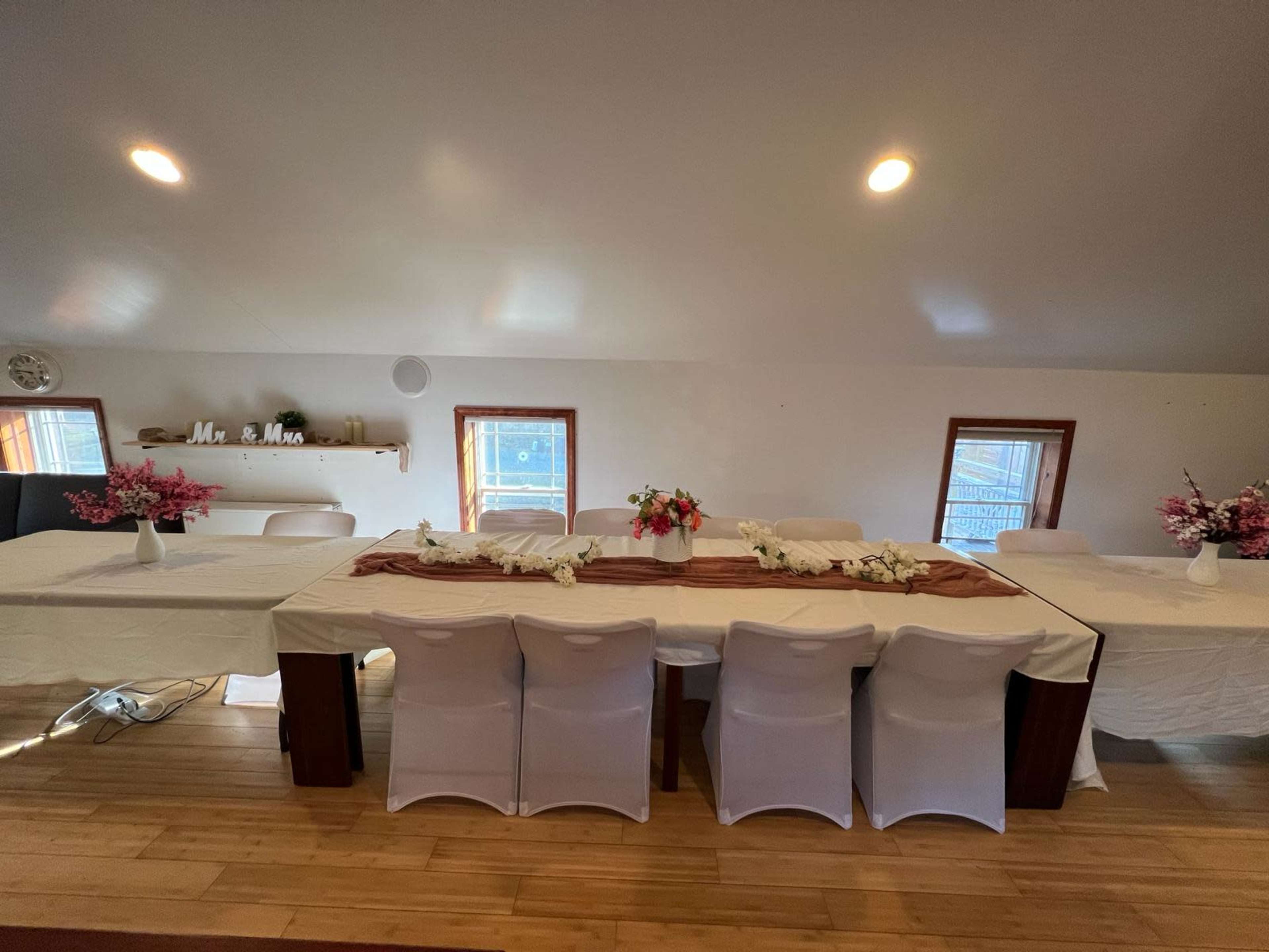 A large table is set for an event with white tablecloths, floral centerpieces, and chairs arranged around it in a bright, open room.