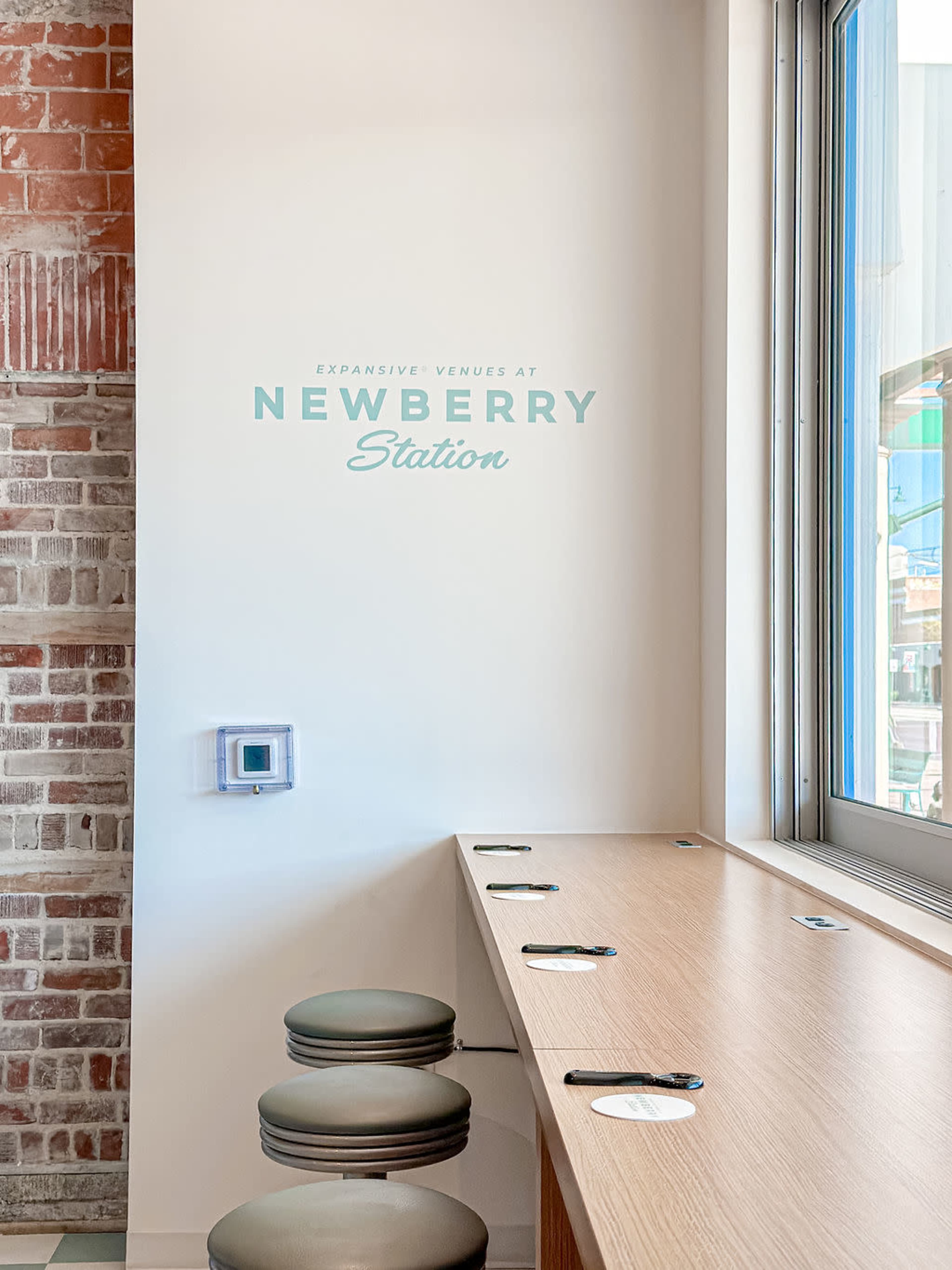 The image shows a modern interior with a wooden counter featuring round seating and a wall displaying the text "Expansive Venues at Newberry Station."