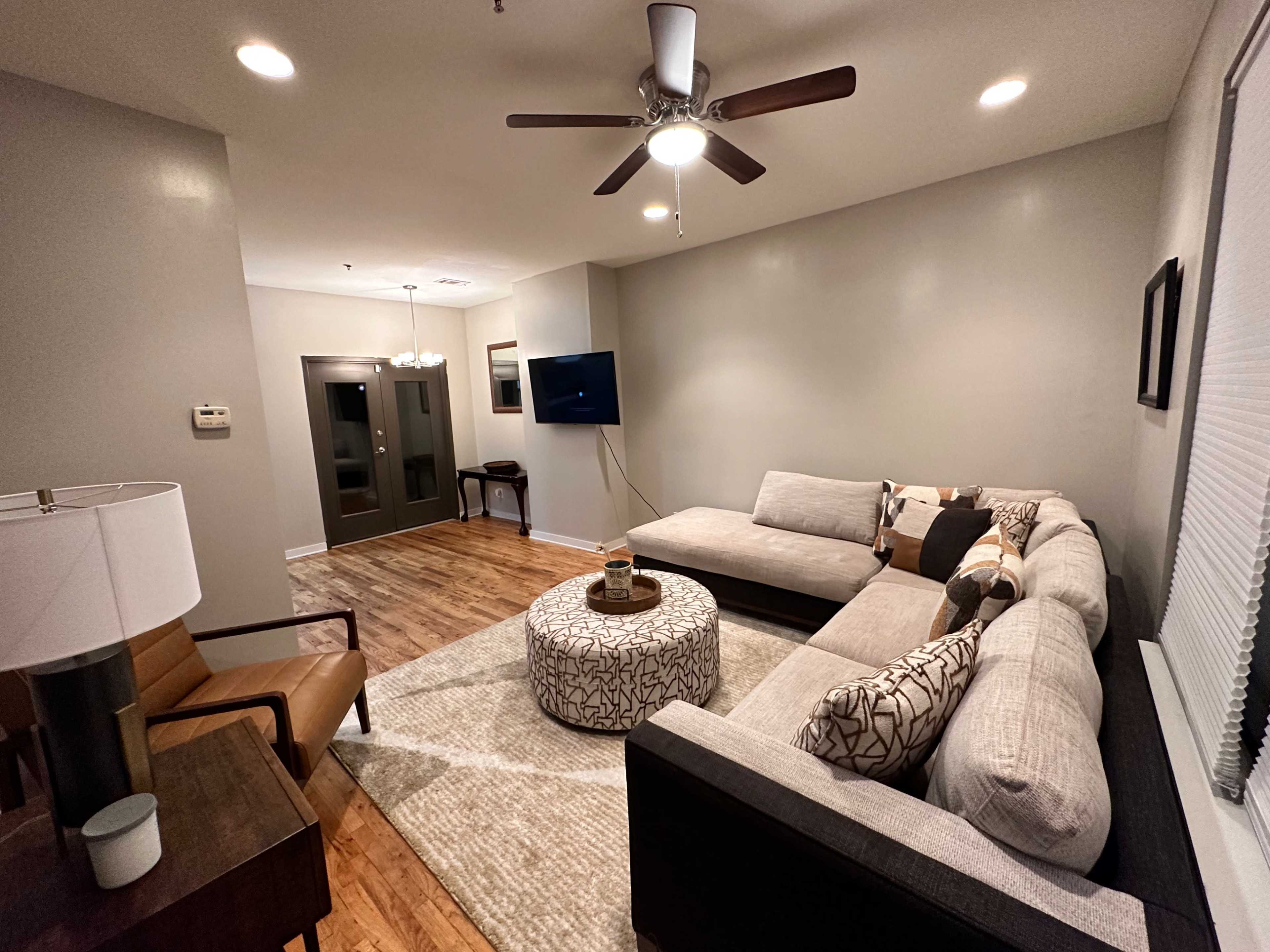 A modern living room features a sectional sofa, round coffee table, and a ceiling fan, with a television mounted on the wall.