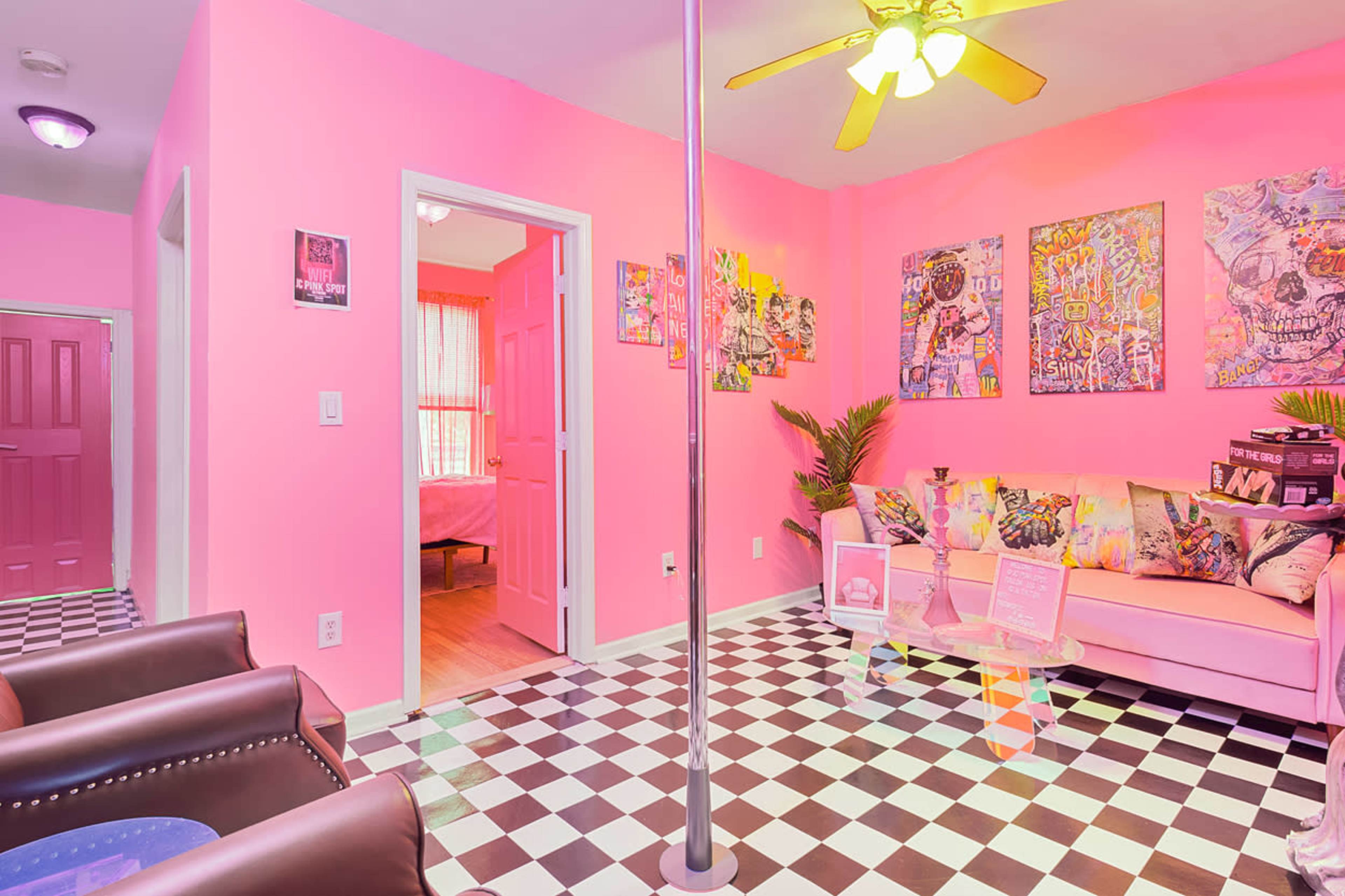 The room features pink walls, black and white checkered flooring, a seating area with colorful cushions, and a pole in the center.