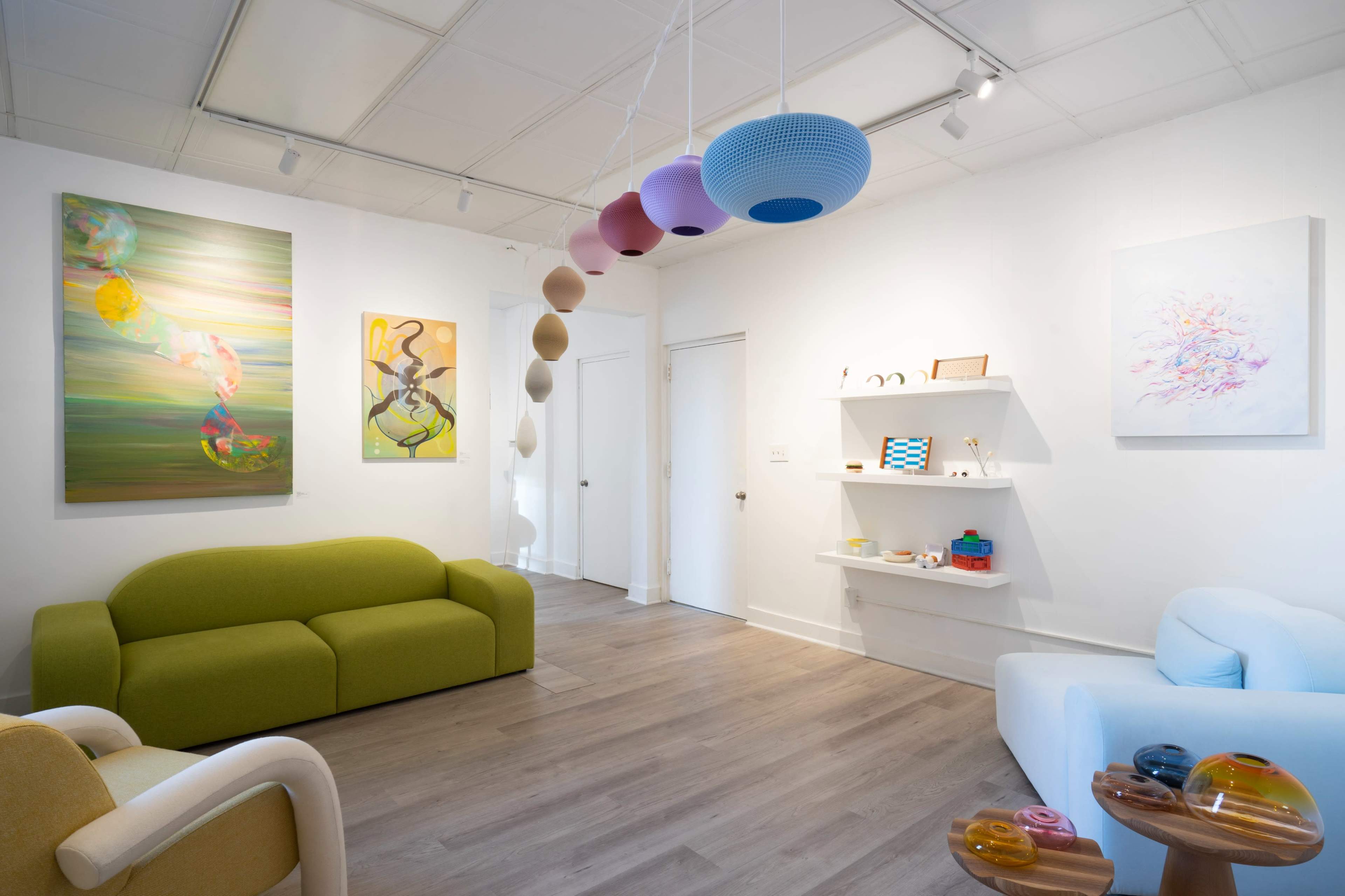 The image shows a modern art gallery with colorful abstract paintings on the walls and minimalist furniture, including a green sofa and a light blue armchair.