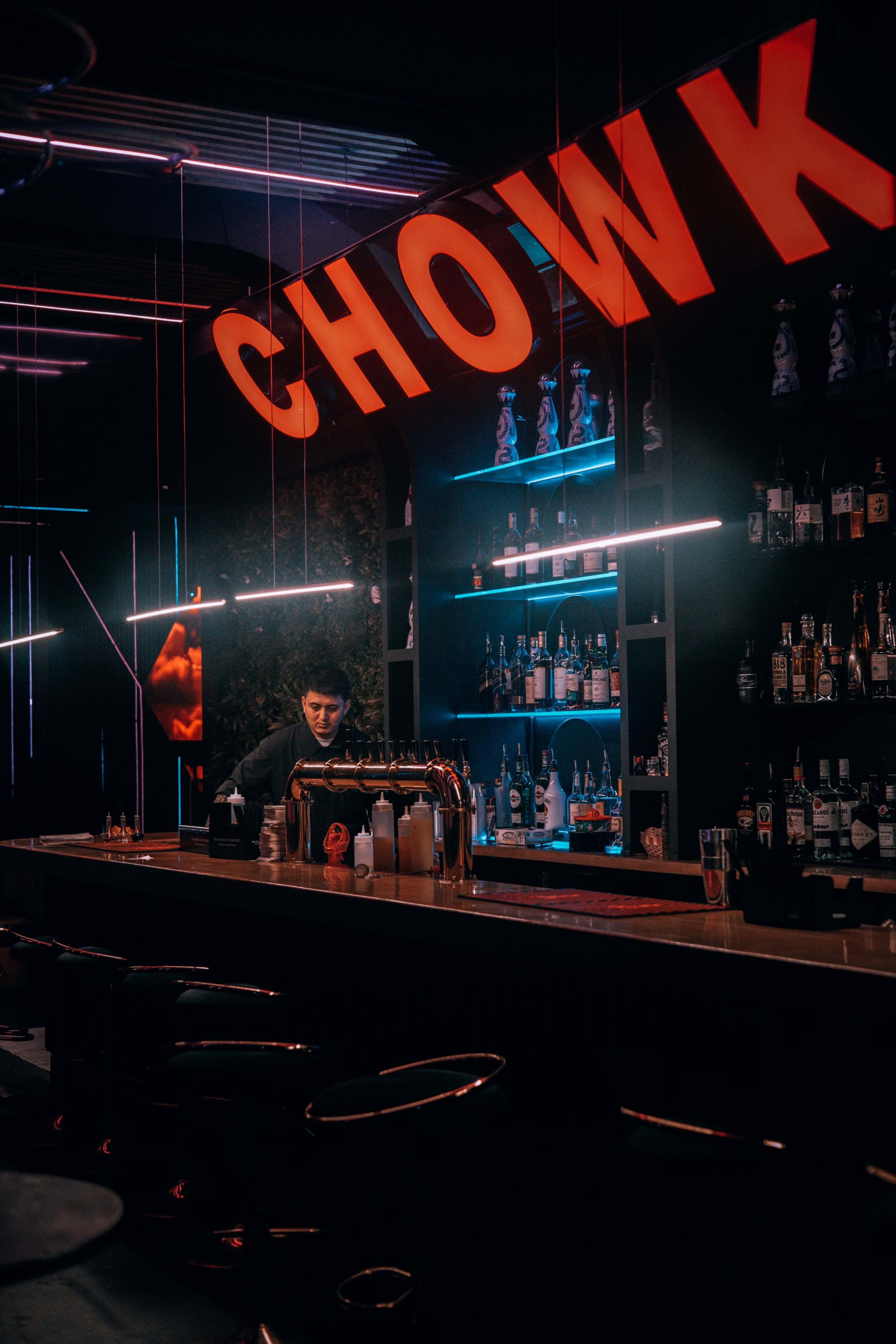 A bartender prepares drinks behind a bar illuminated by red neon signs in a dimly lit space.