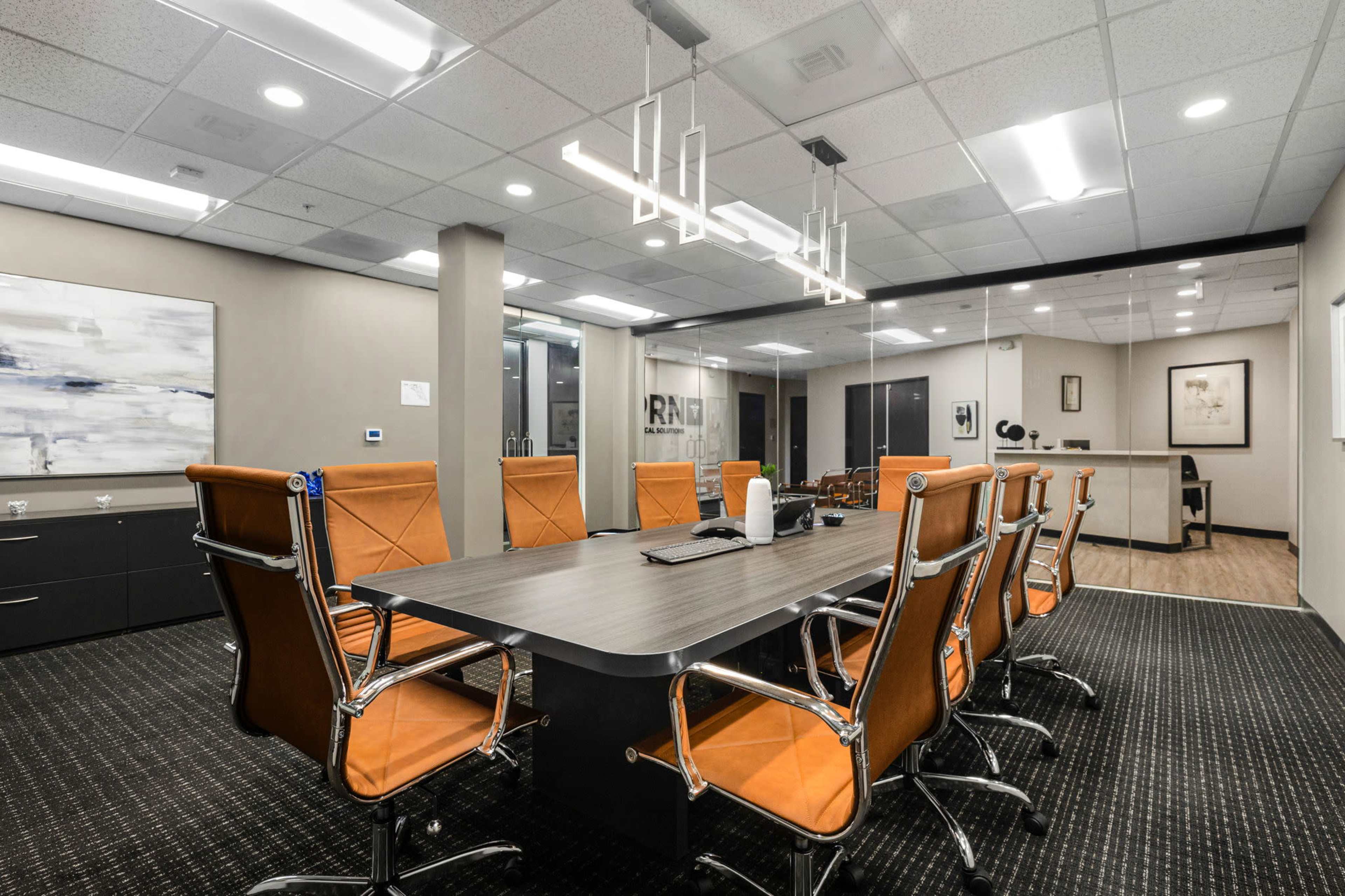 A modern conference room features a long table surrounded by orange office chairs and is equipped with overhead lighting and glass walls.