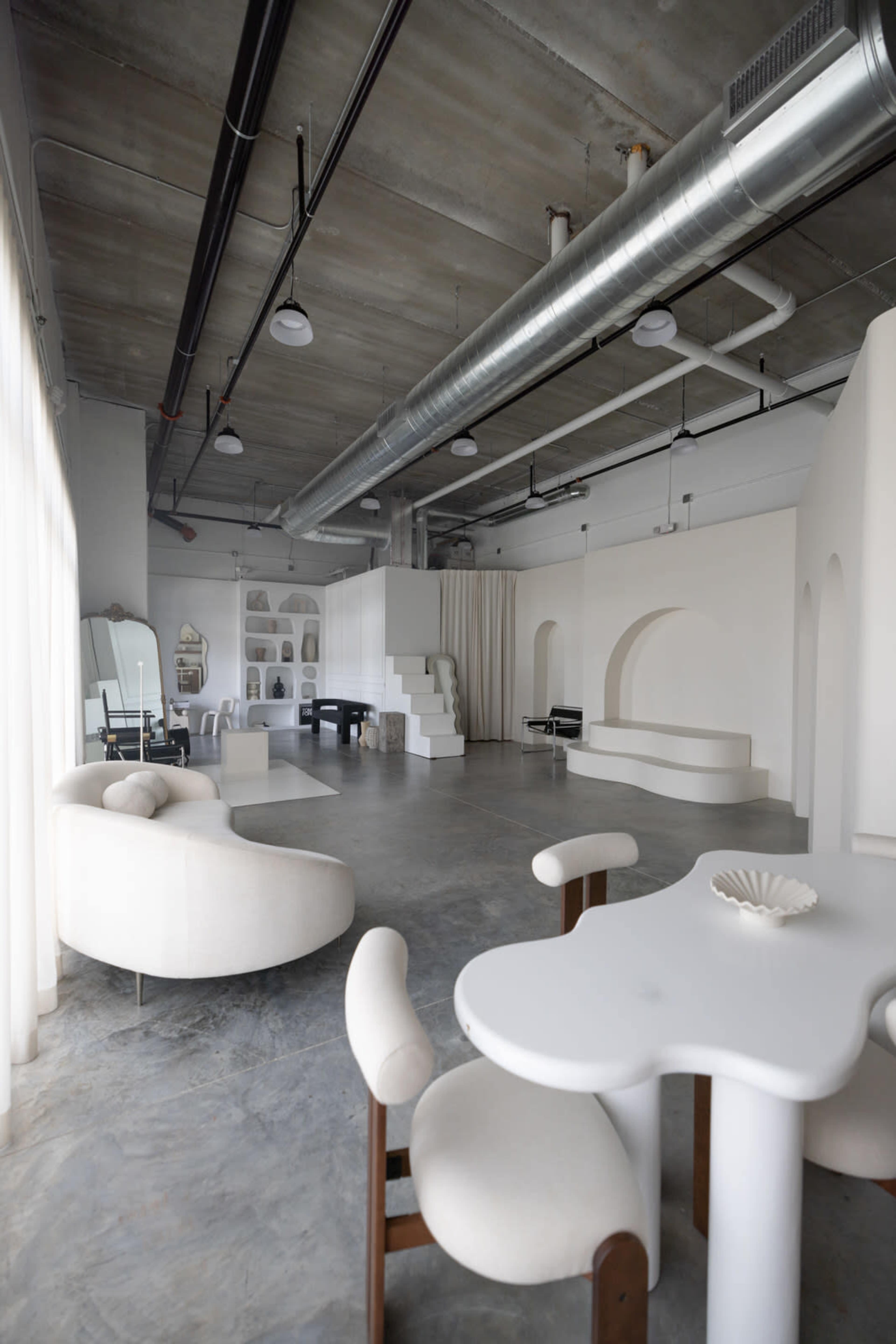 A modern interior space with minimalist furniture, including a curved sofa and a unique table, set against a backdrop of white walls and concrete flooring.