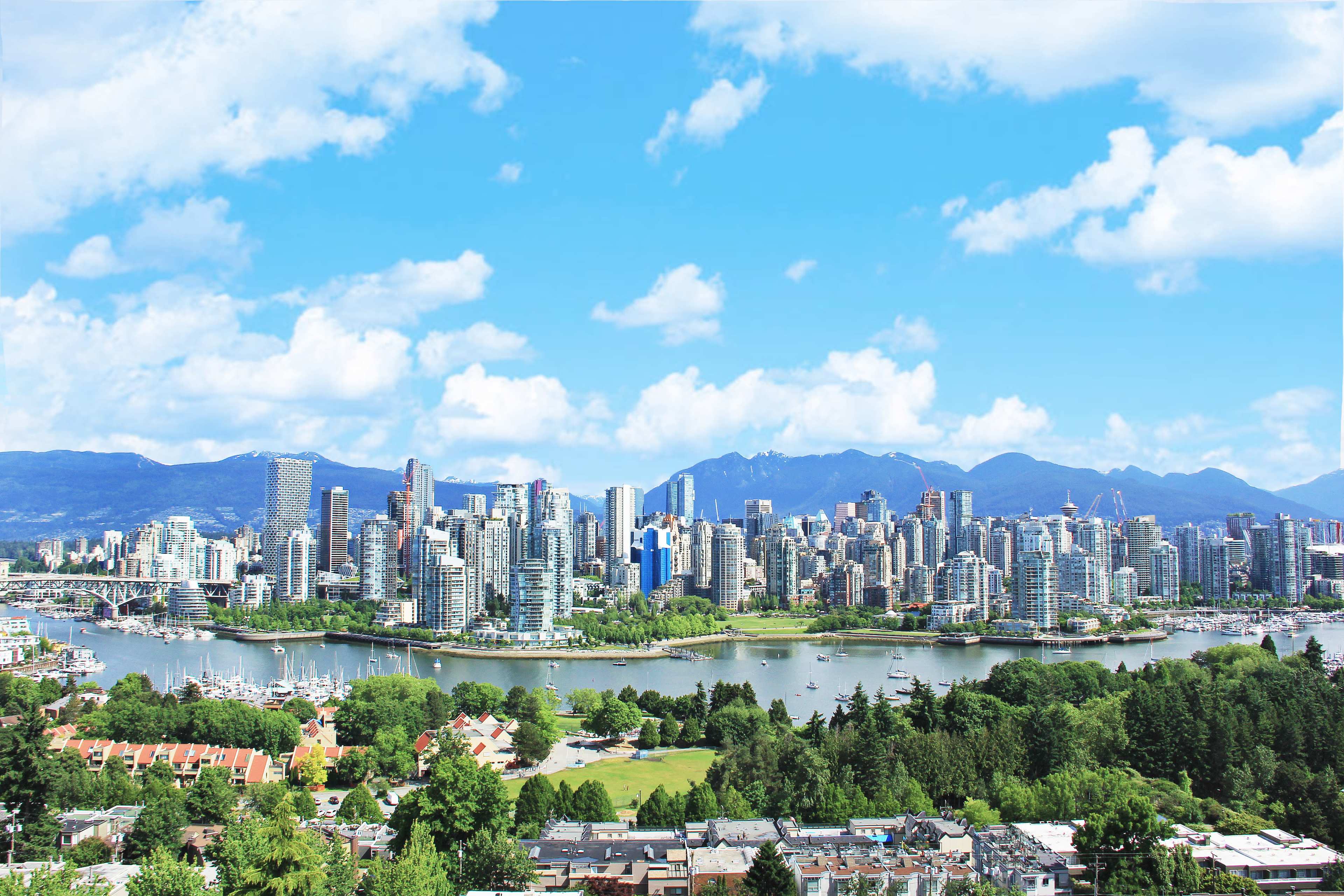 A panoramic view of Vancouver showcases its modern skyline alongside lush greenery and mountains under a clear blue sky.