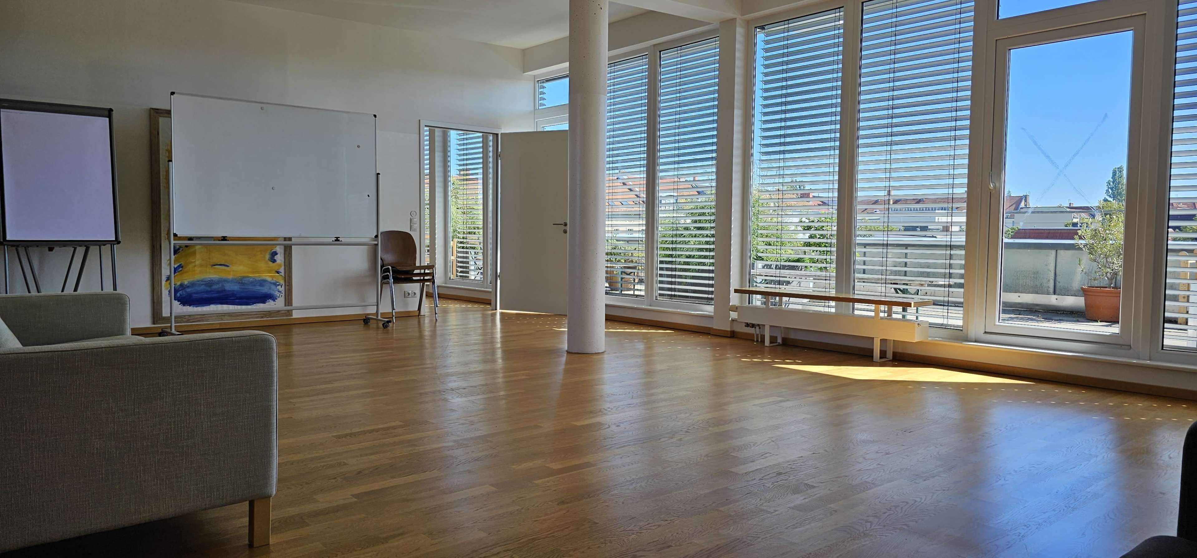 A bright and spacious room features large windows with blinds, a whiteboard, and a chair, complemented by wooden flooring.