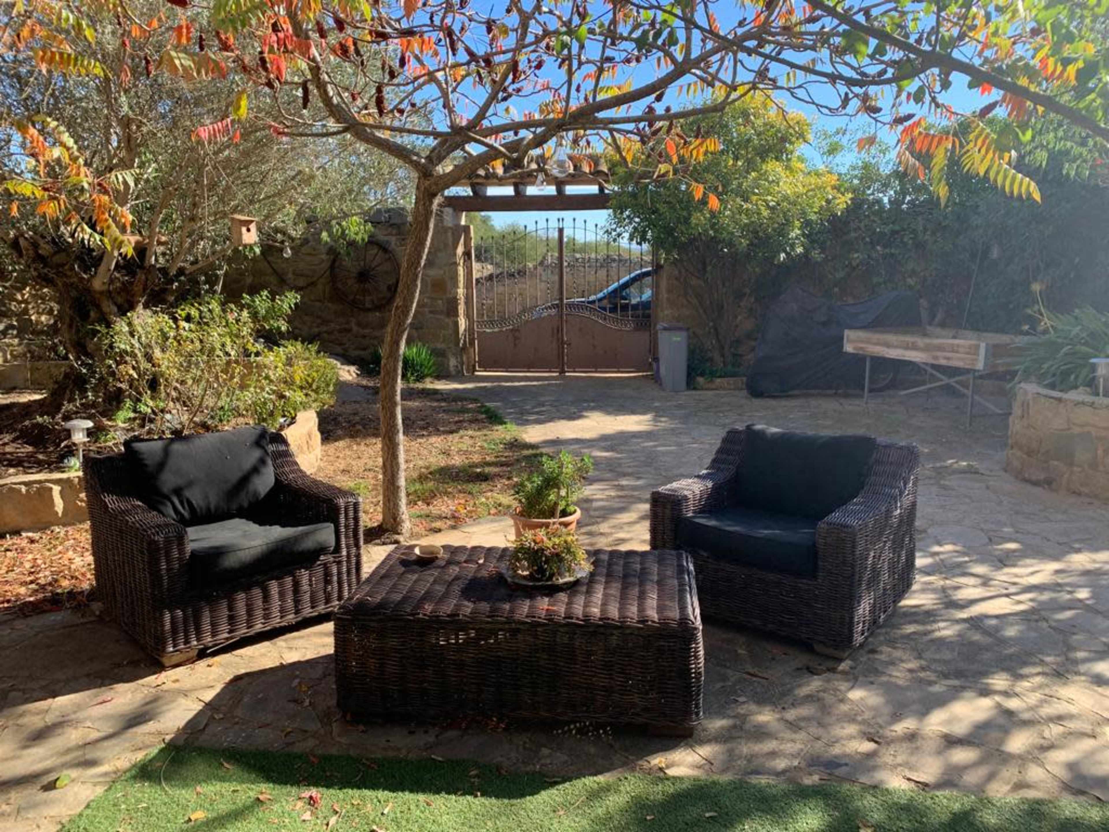 A patio area features two wicker chairs and a table surrounded by trees and a gate in the background.
