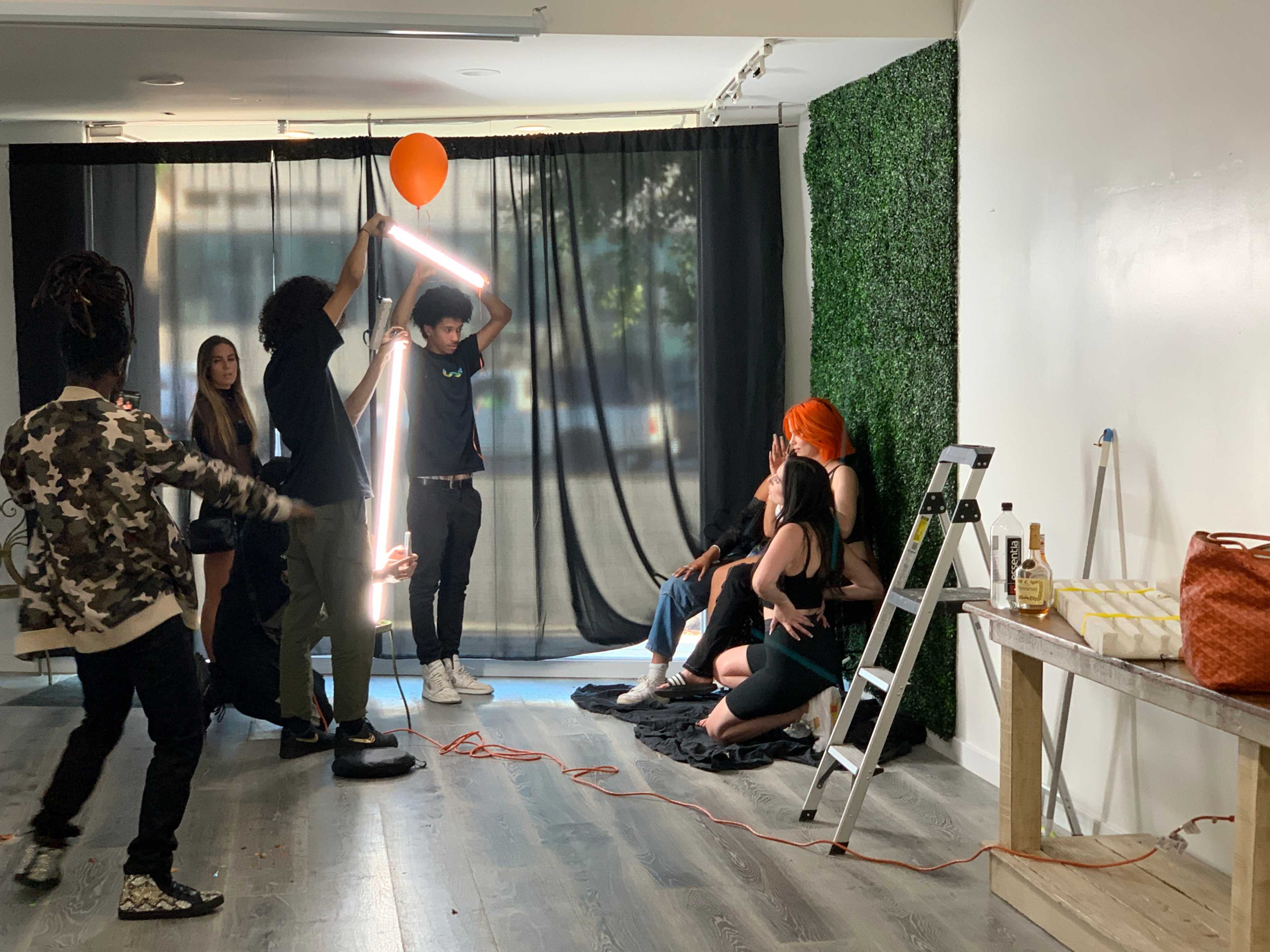 A group of people poses for a photo shoot in a studio, with a person holding a neon light and another holding a balloon.