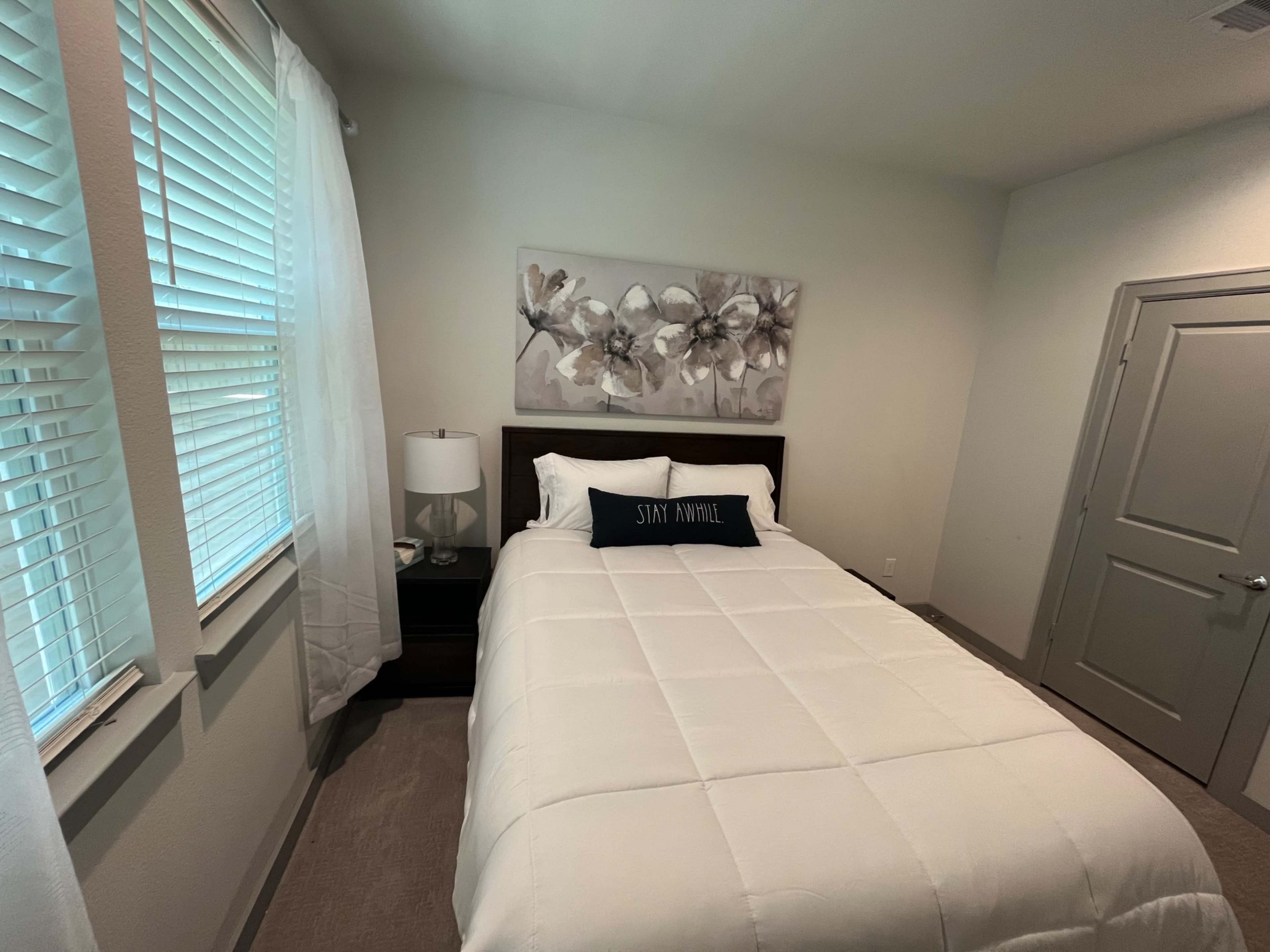 A neatly arranged bedroom featuring a bed with a white comforter, a bedside table with a lamp, and a piece of floral artwork on the wall.