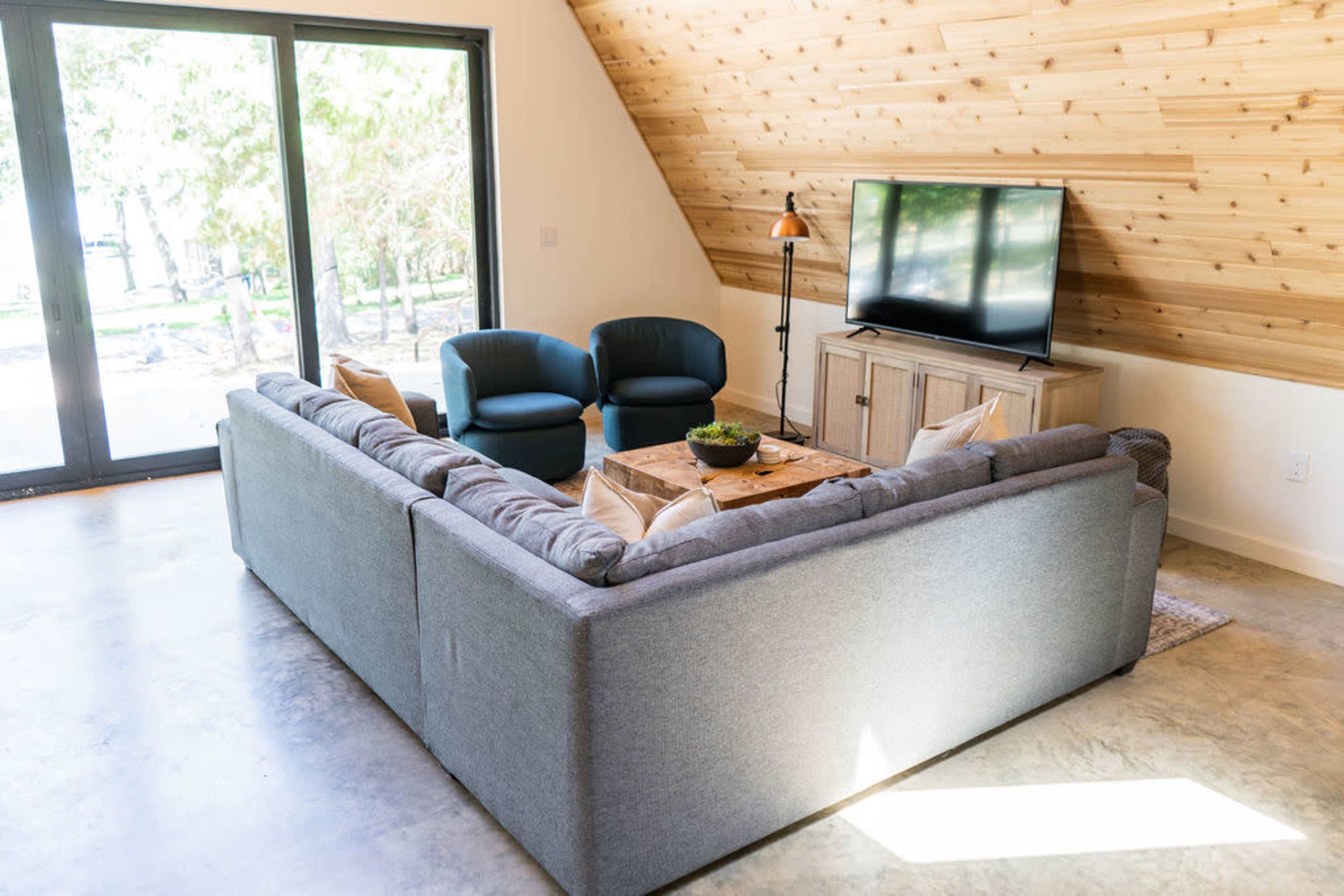 A cozy living room features a large gray sectional sofa, two blue armchairs, a coffee table, and a television against a wood-paneled wall.