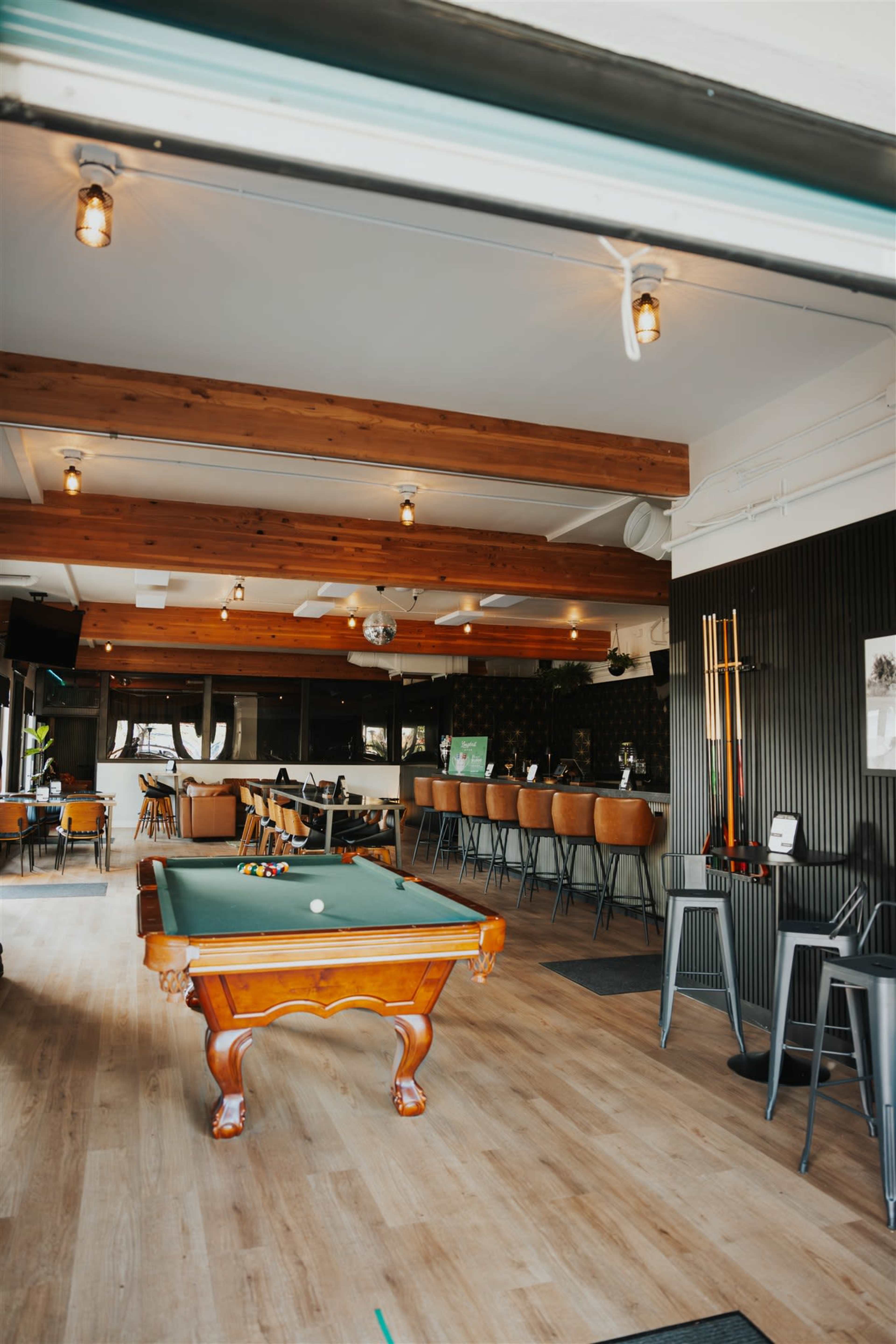 The image shows a spacious bar area featuring a wooden pool table, seating at a bar counter, and modern decor with pendant lighting.