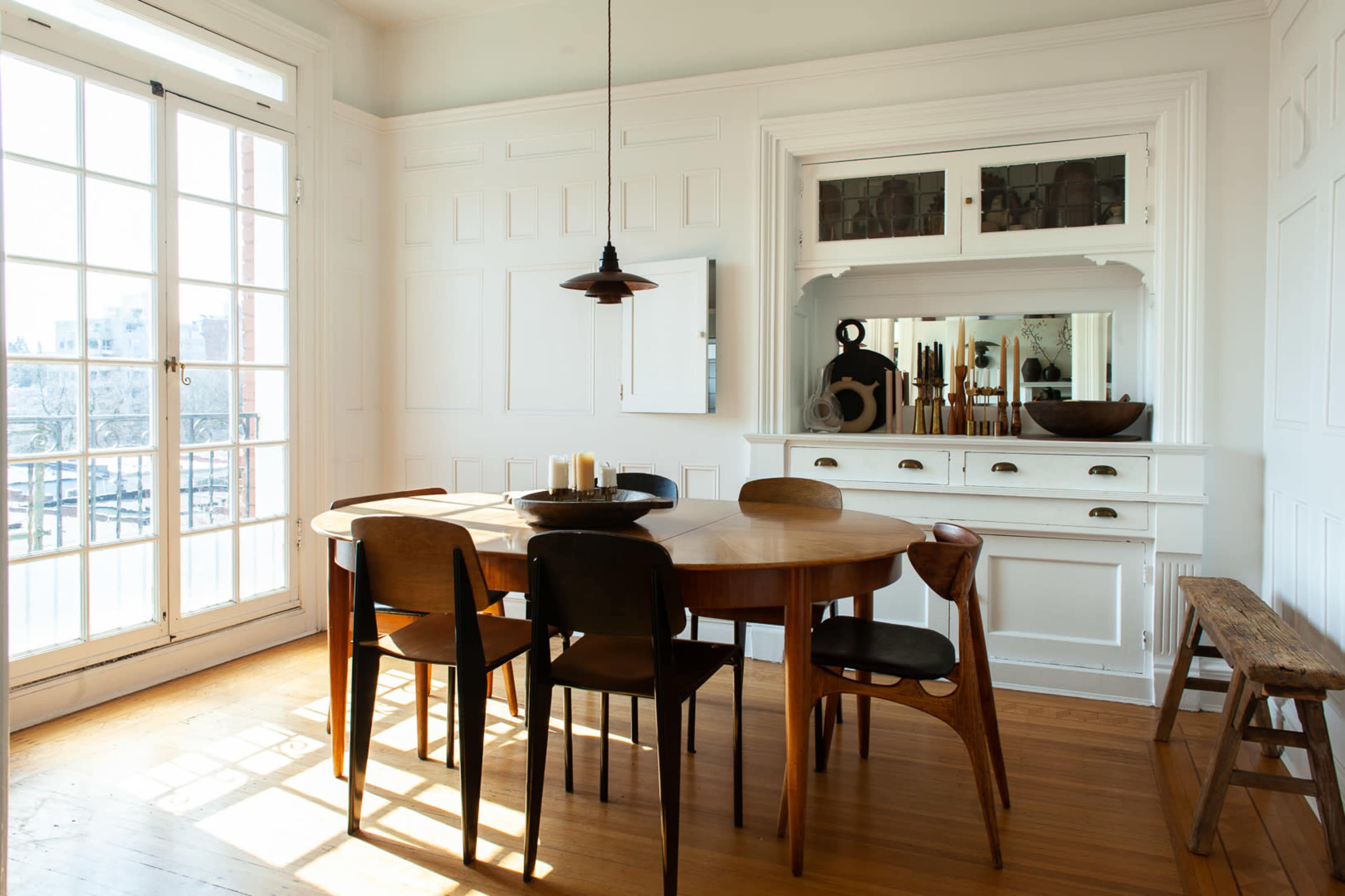 A dining room features a round wooden table surrounded by six chairs, with a minimalist sideboard and large windows allowing natural light to enter.