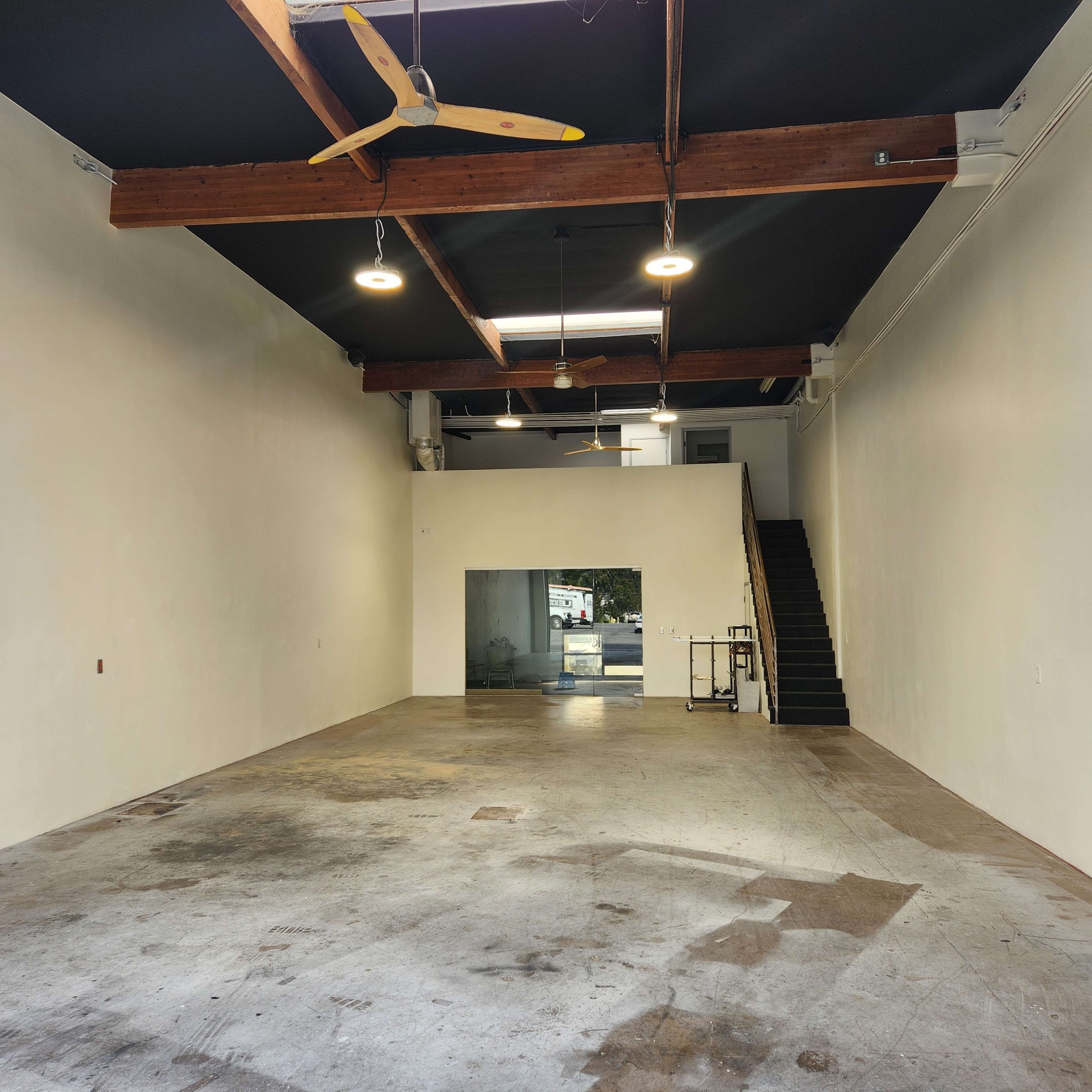 An empty interior space with exposed wooden beams, ceiling fans, and a staircase leading to an upper level.