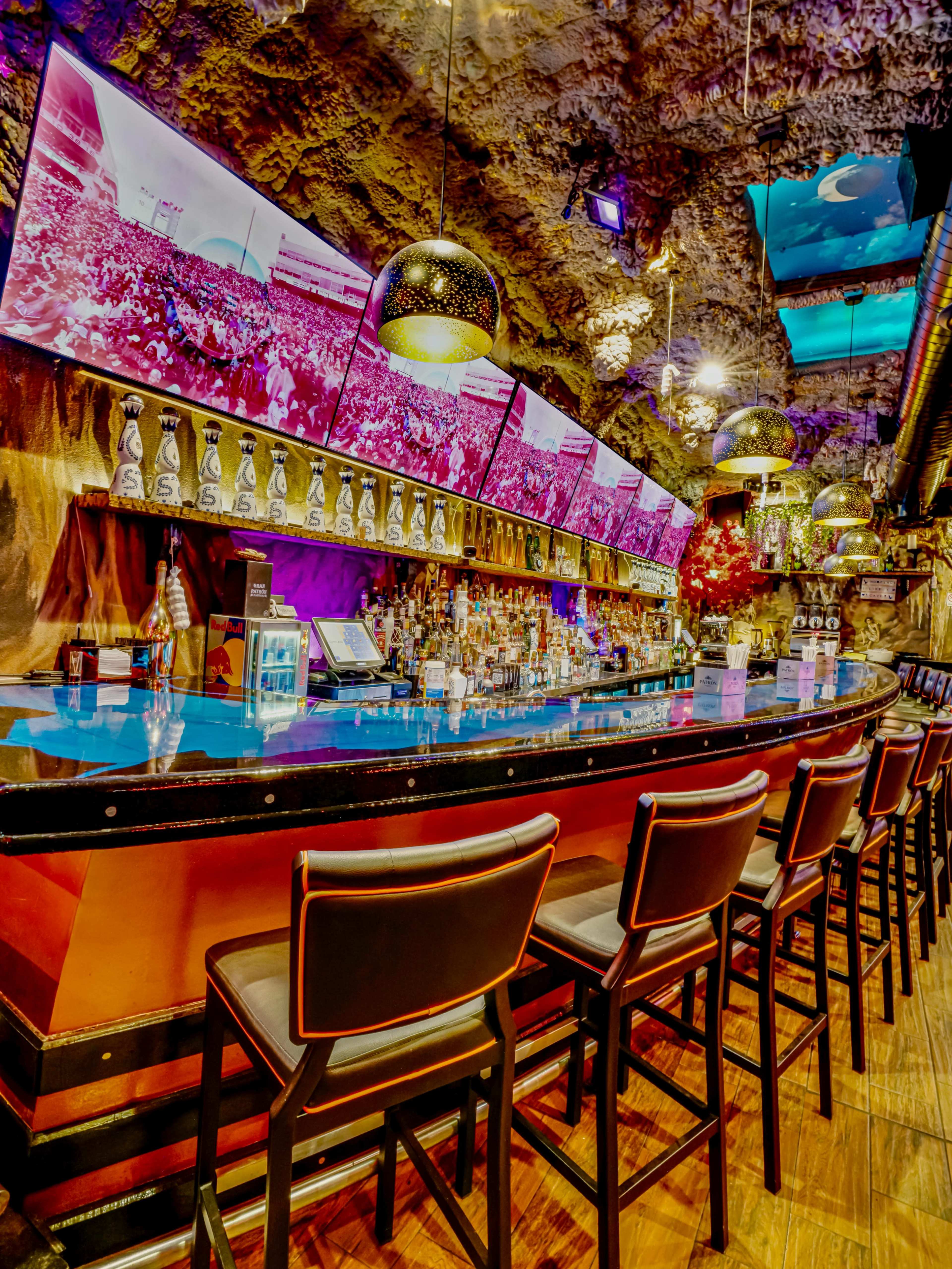 The image shows a well-lit bar featuring a curved counter, high stools, and several large screens displaying vibrant images in a cave-like setting.