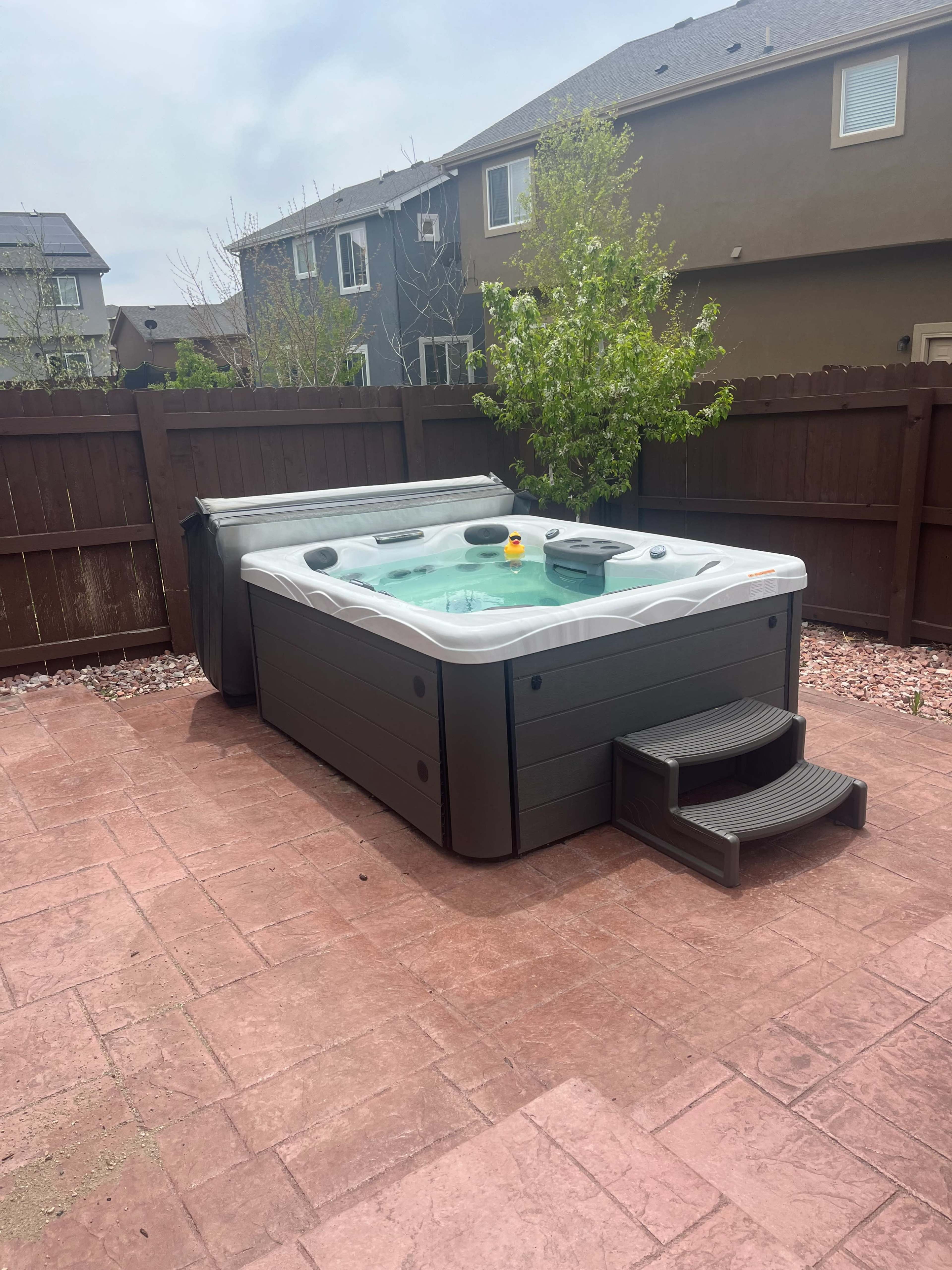 The image shows a hot tub surrounded by a patio made of pavers, with a fenced backyard in the background and a few residential buildings visible.