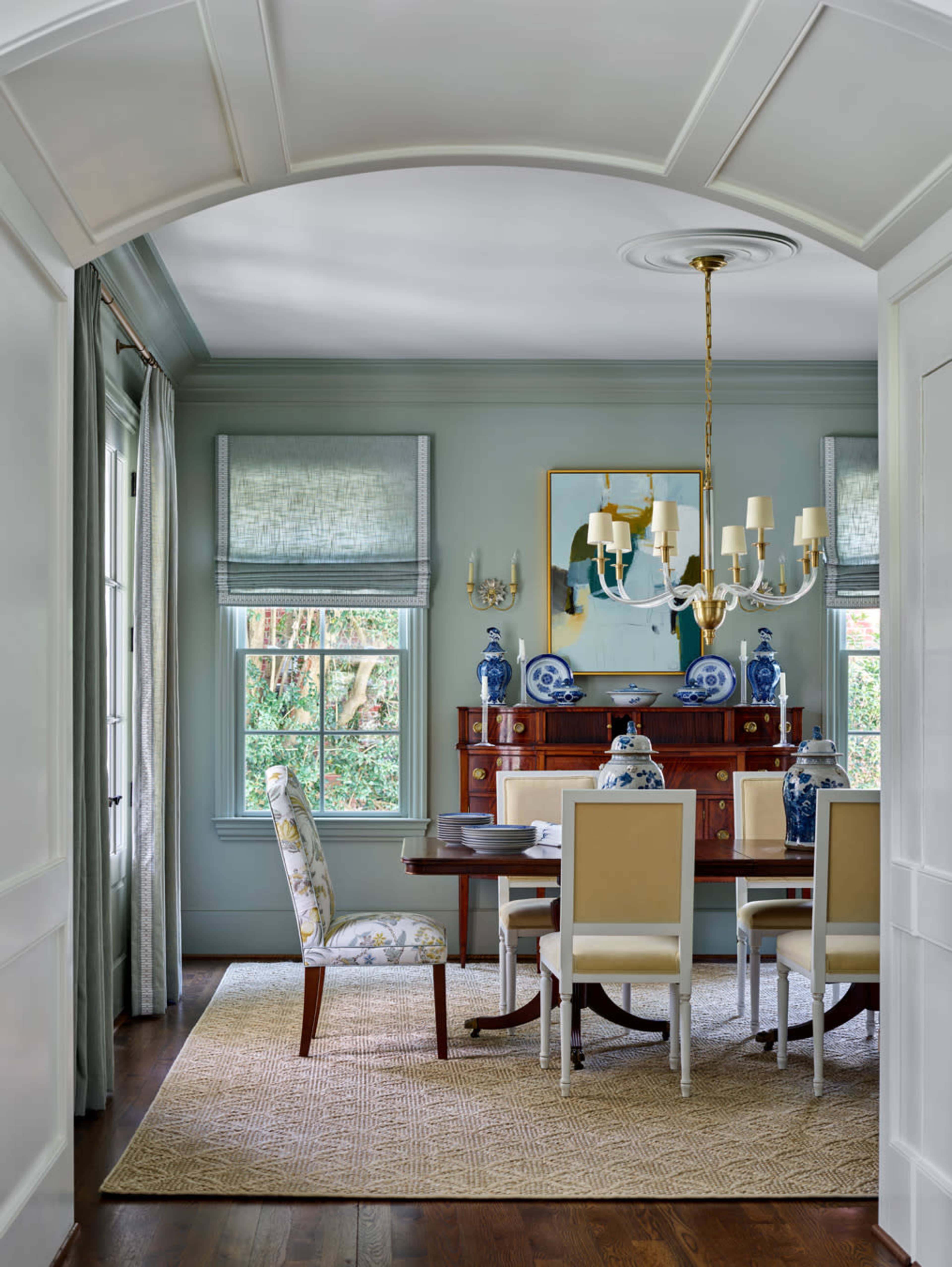 A dining room features a wooden table surrounded by upholstered chairs, with a sideboard displaying blue and white decor, and large windows letting in natural light.