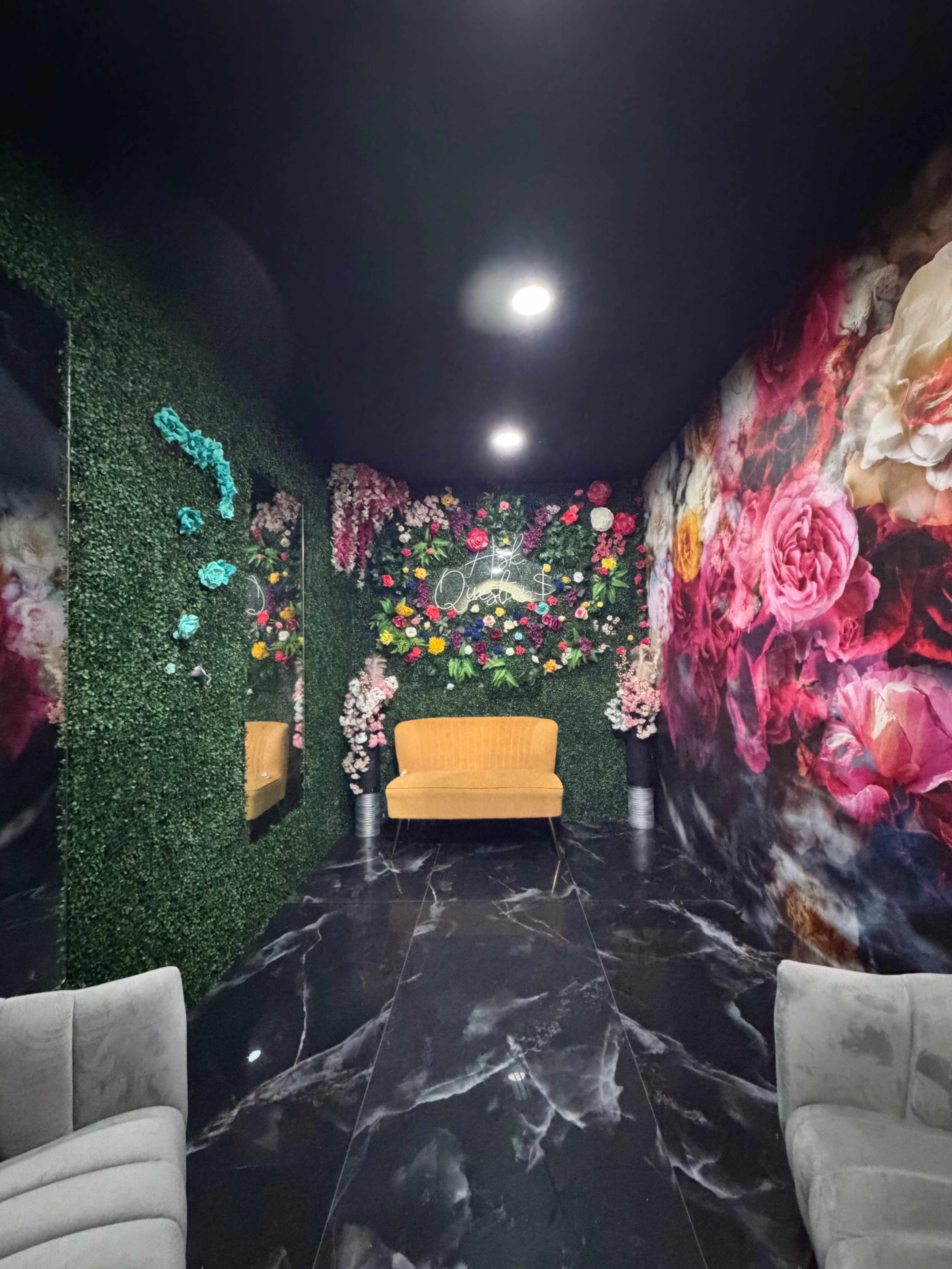 The image depicts a small, decorated room featuring artificial greenery and floral wall art, with an orange seat at the center and mirrors along the walls.