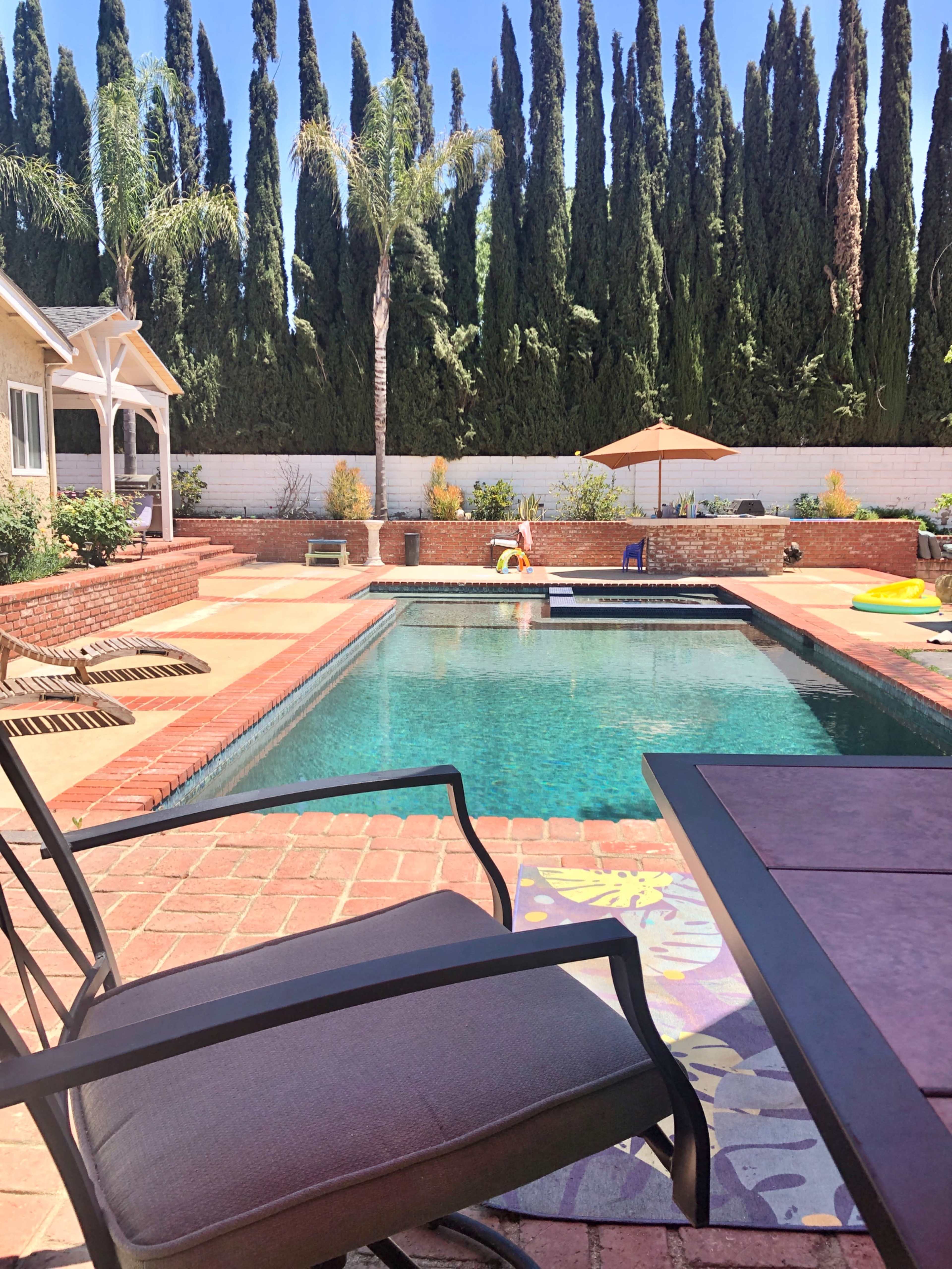 A swimming pool with a brick patio is surrounded by tall green trees and a covered seating area.