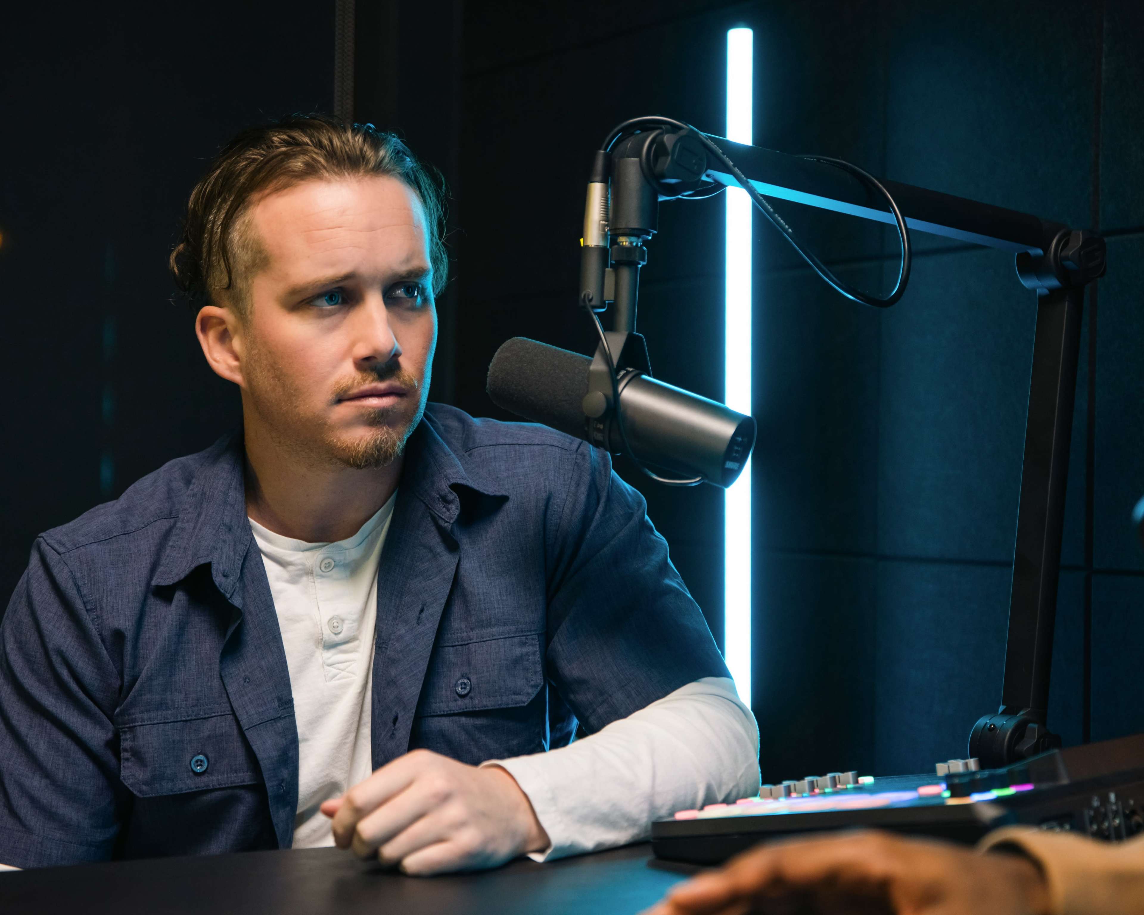 A man with medium-length hair sits at a microphone in a recording studio, observing intently.