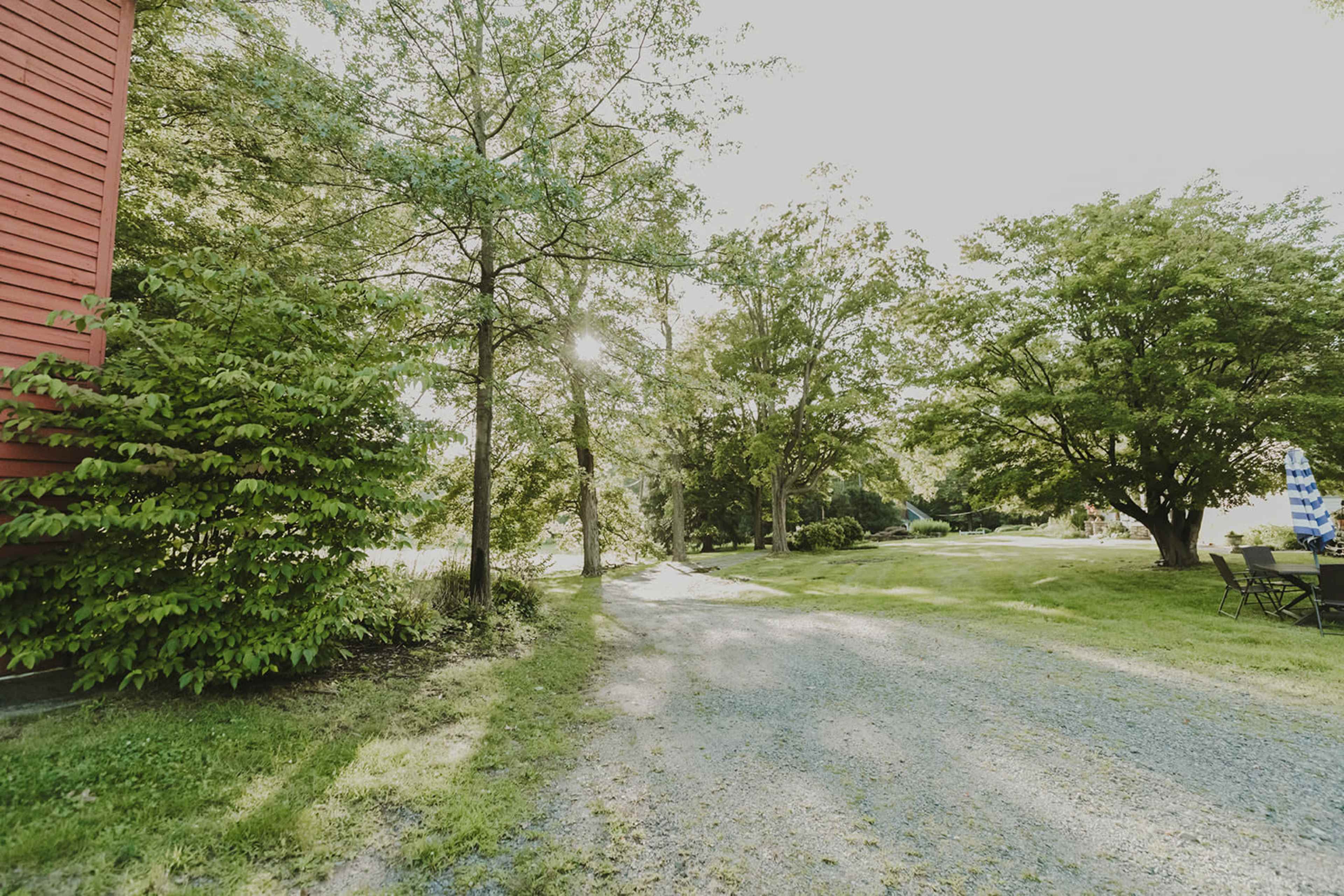 A gravel path leads through green grass and trees beside a red building, with sunlight filtering through the leaves.