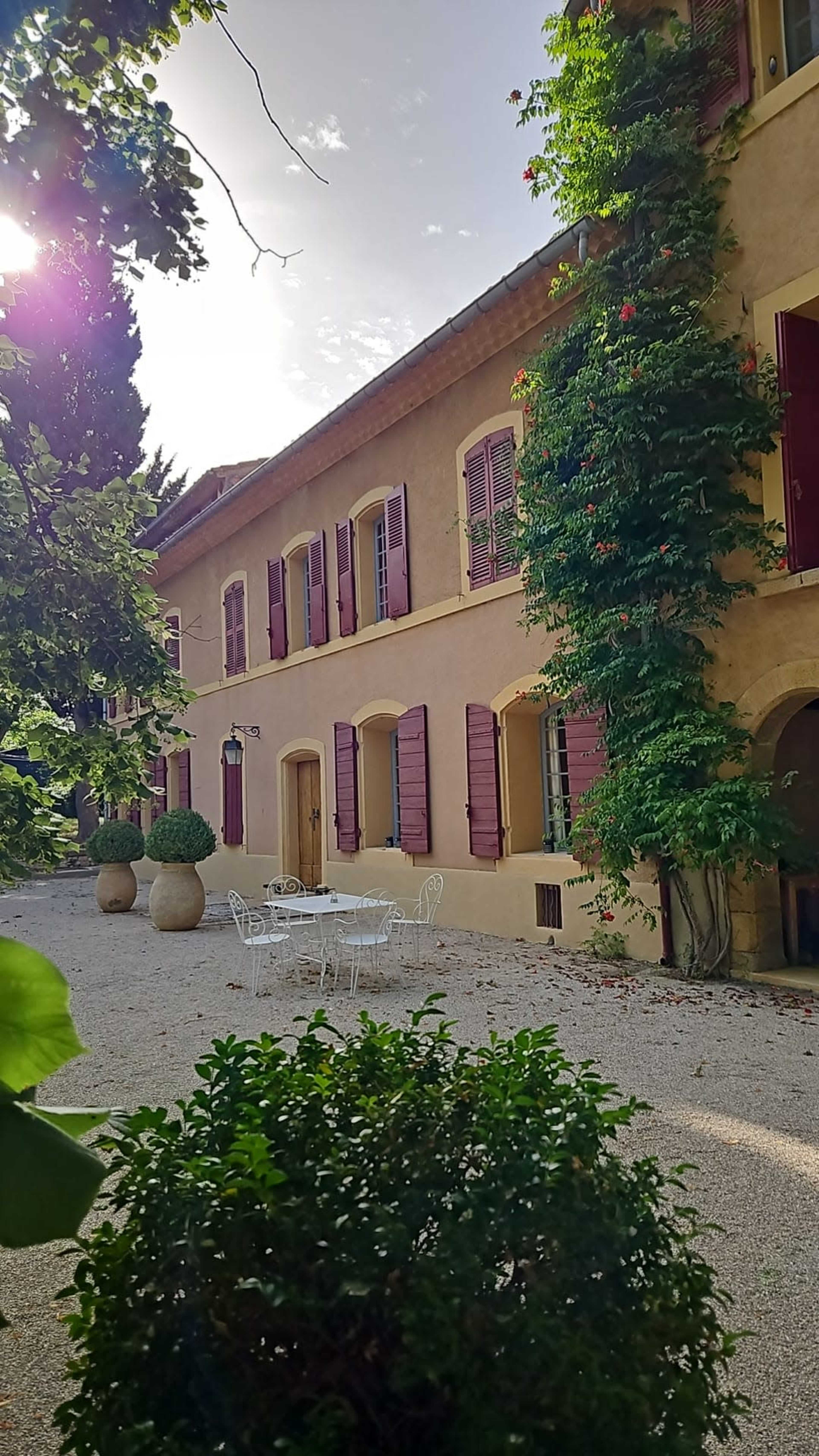A sunlight-drenched building with shutters and a vine-covered wall is surrounded by a gravel courtyard featuring a small white table and chairs.