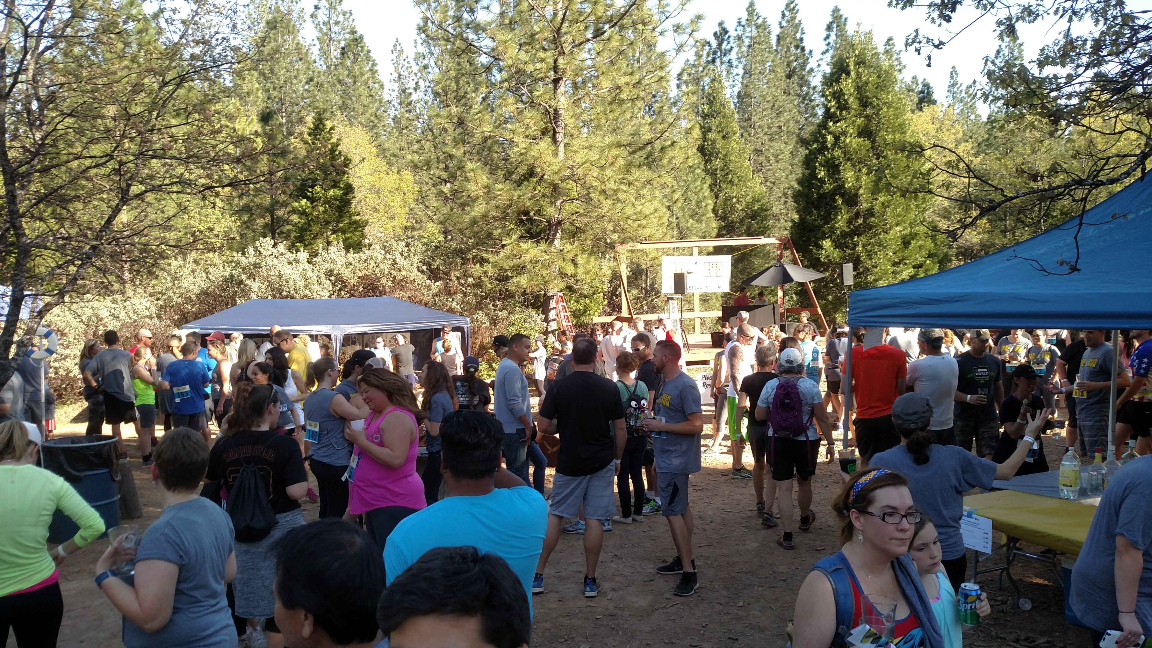 A large group of people gathers in a forested area for an outdoor event, with tents and a stage visible in the background.