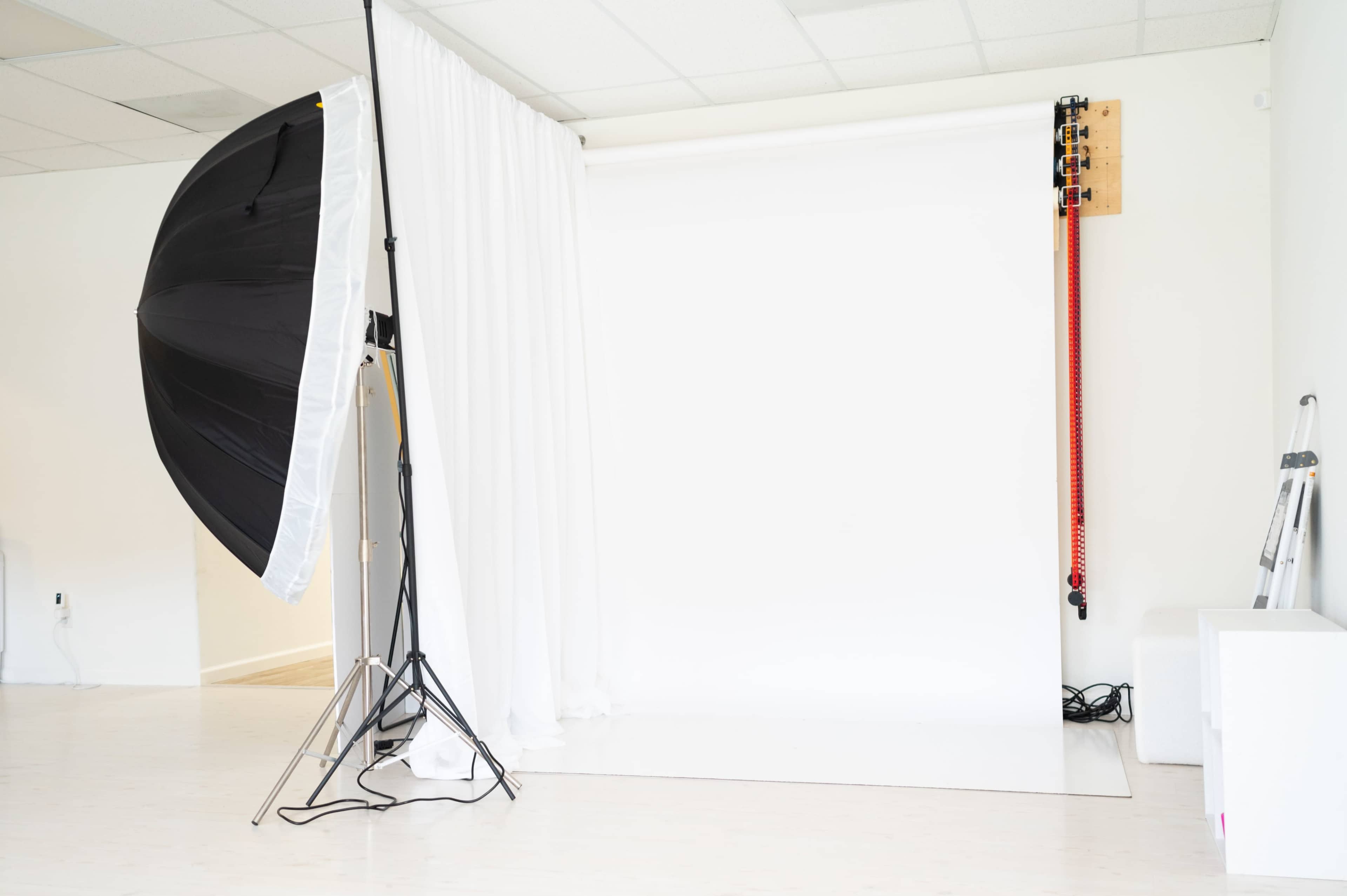 A photography studio features a large softbox light, a white backdrop, and a ladder against a plain white wall.