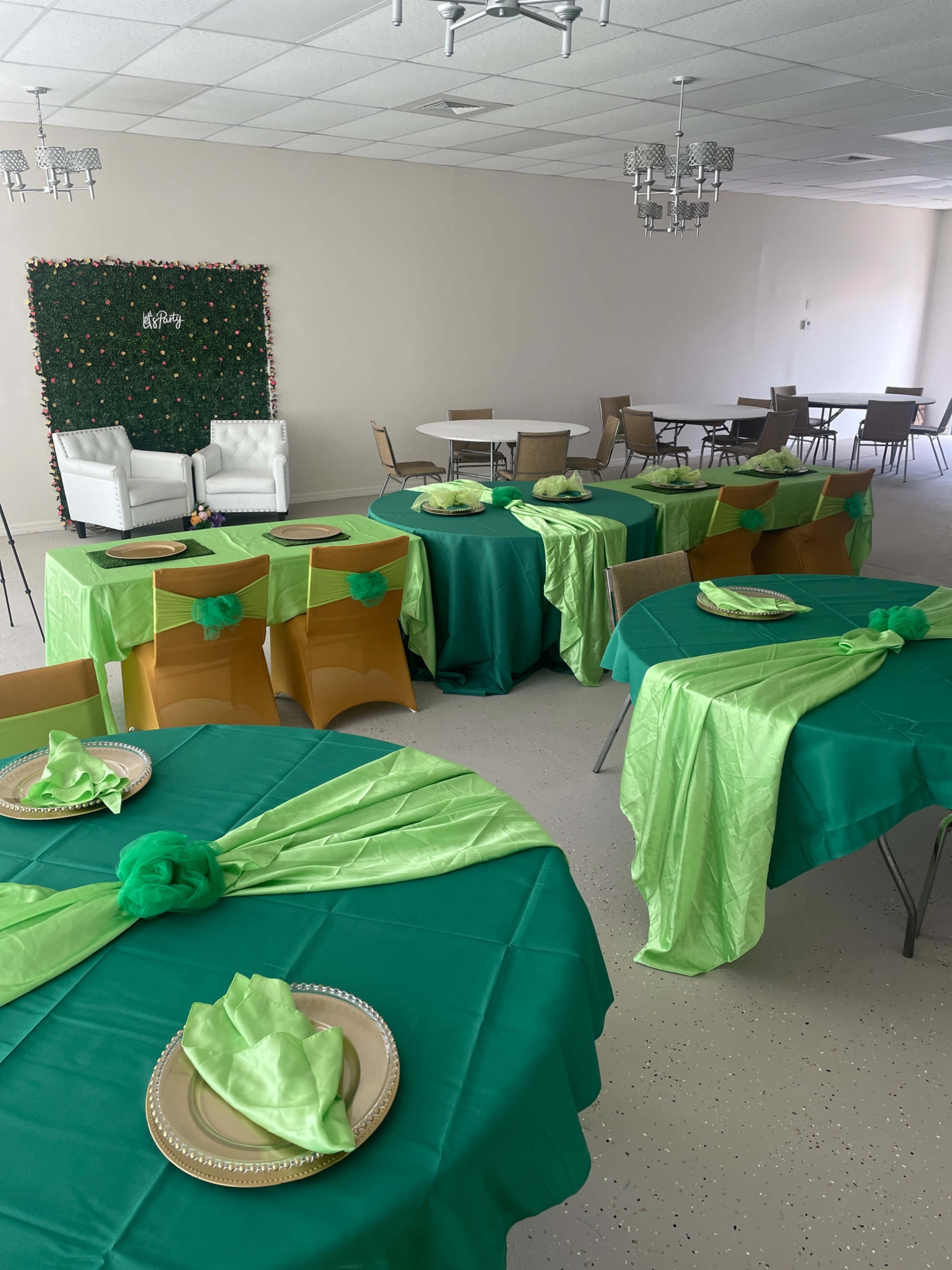 A banquet setup with green and gold tablecloths arranged on multiple tables, featuring plates and decorative accents, in a room with additional seating and a green backdrop.