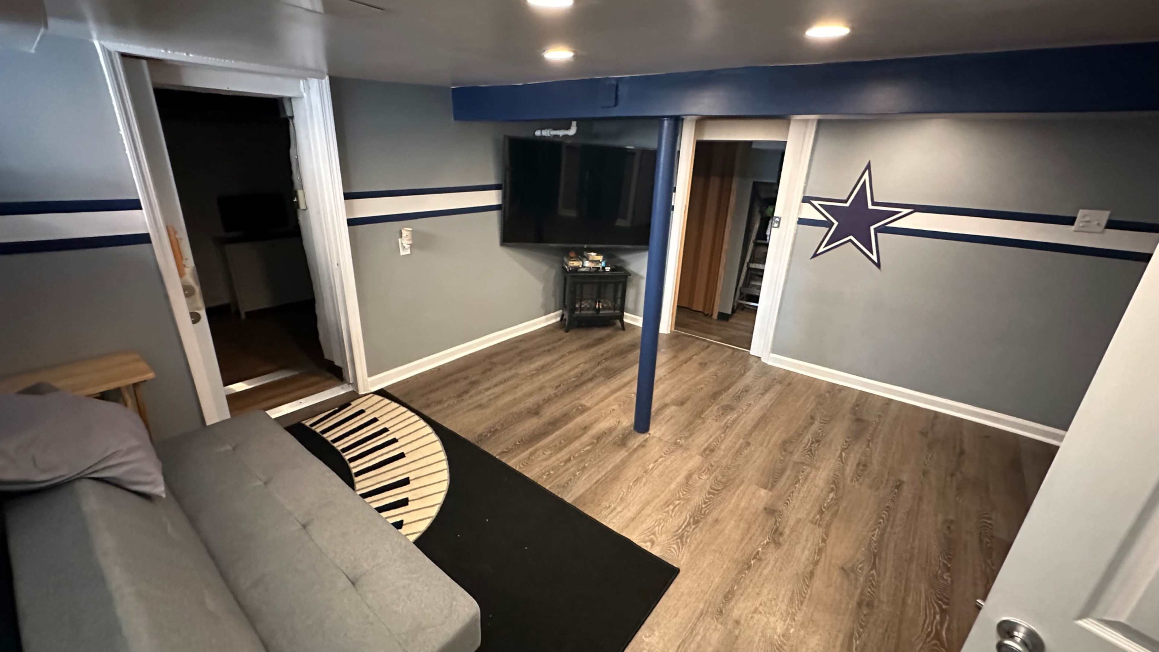 A basement room with a gray sofa, a black rug shaped like piano keys, a television mounted on the wall, and a star design on the wall.