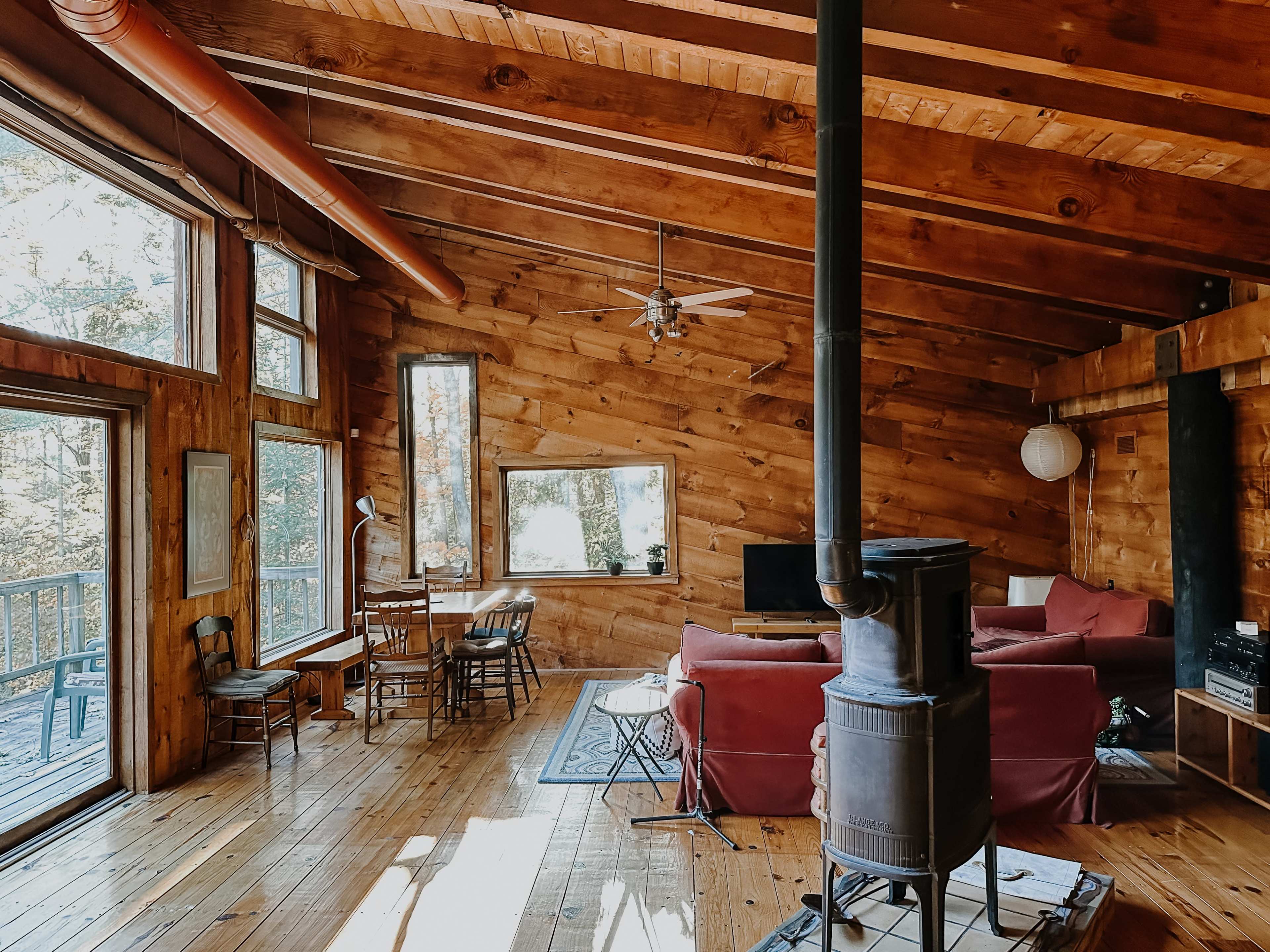 The image shows a wooden cabin interior featuring a living area with a red couch, a dining table, and large windows allowing natural light to enter.