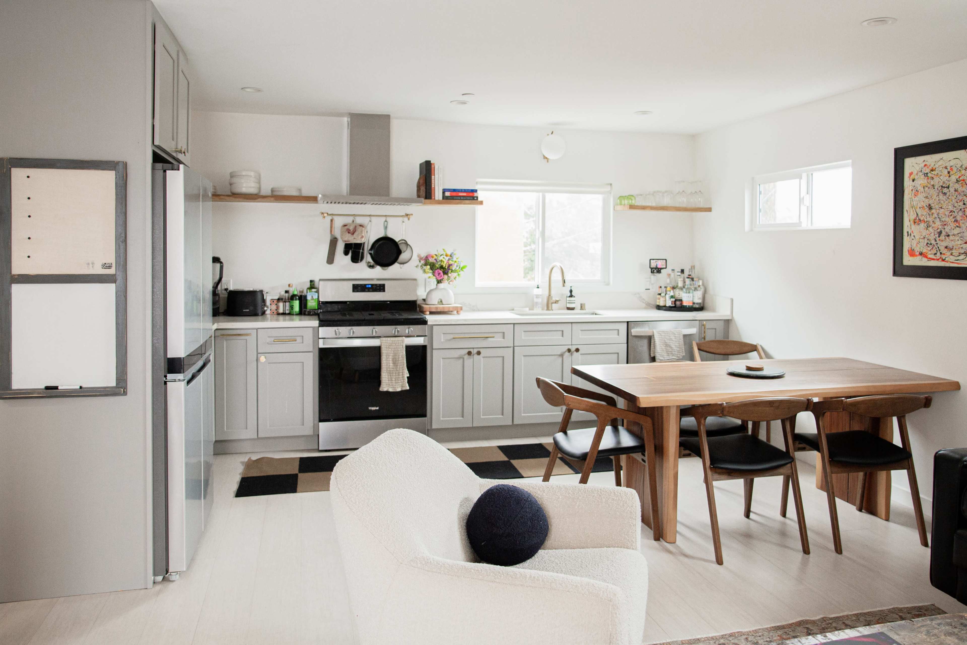 The image shows a modern kitchen with gray cabinetry, stainless steel appliances, a large wooden dining table, and a cozy seating area.
