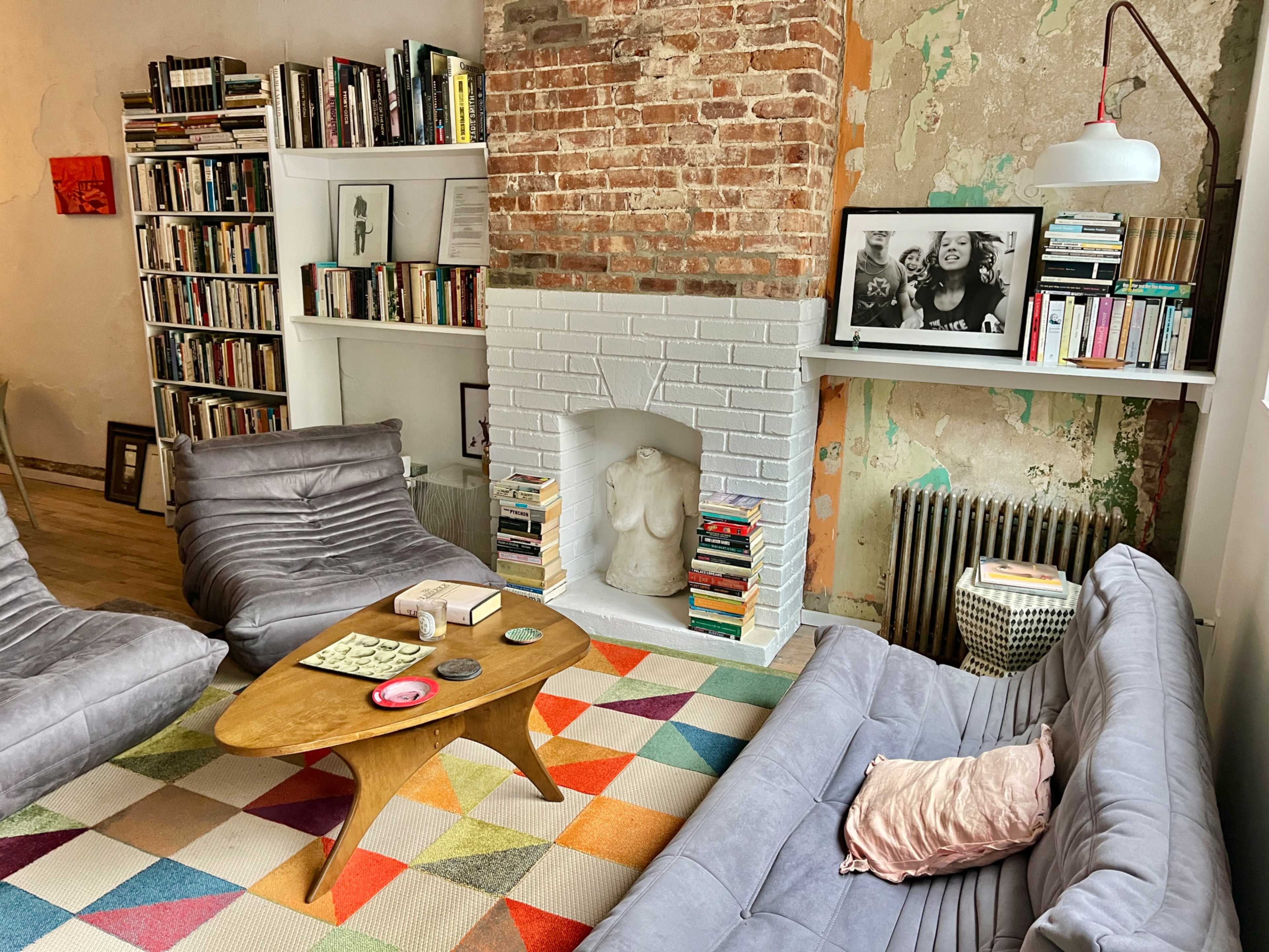 The image shows a cozy living room with two gray lounge chairs, a colorful patterned rug, a wooden coffee table, and bookshelves filled with books along a brick and painted wall.