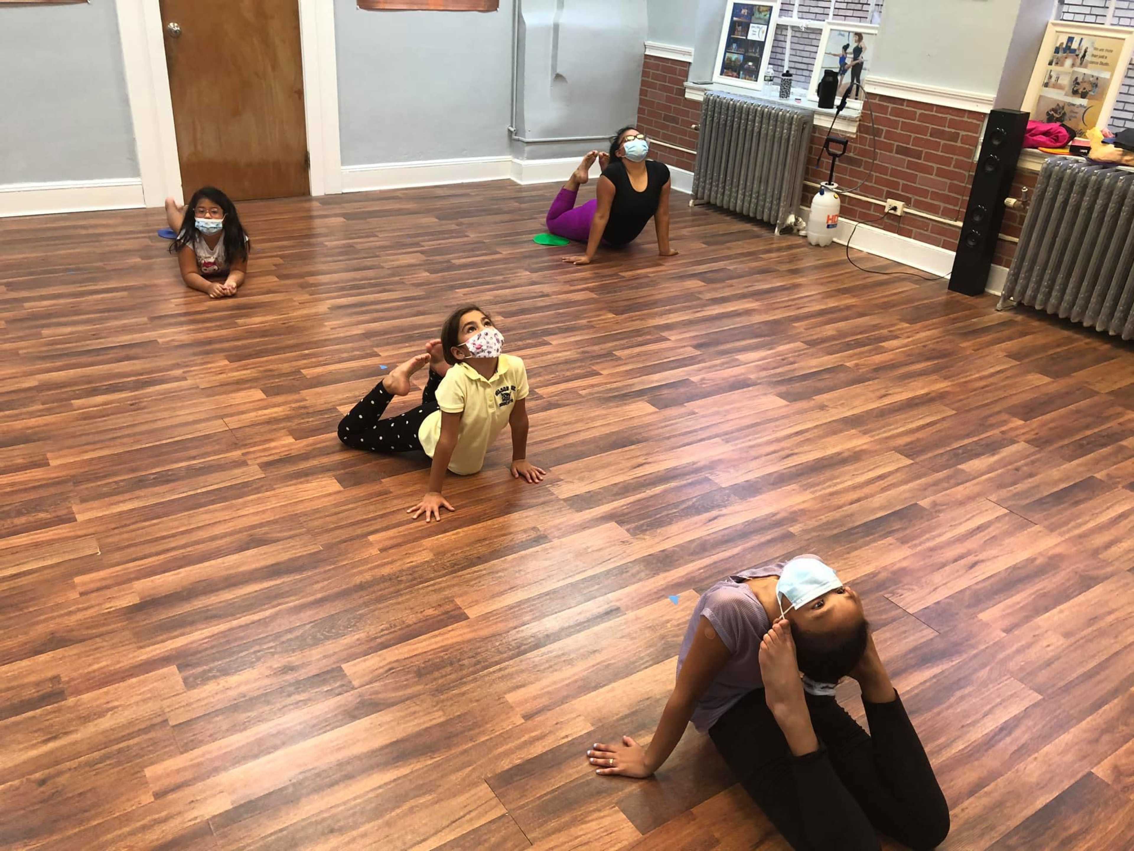 Five individuals are practicing yoga poses on a wooden floor in a studio while wearing masks.