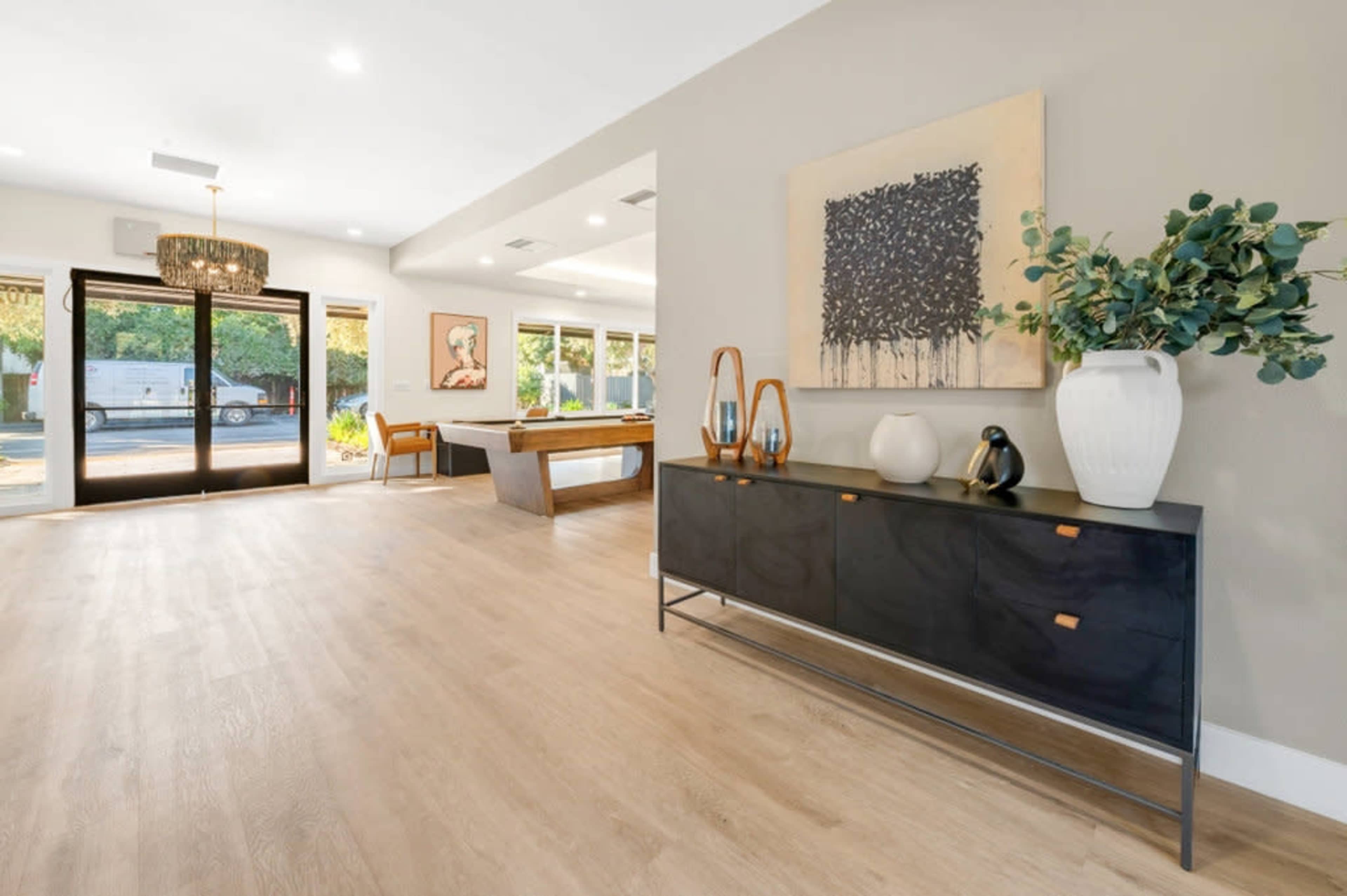 The image shows a spacious, modern living area featuring a black cabinet with decorative items, large windows, and a pool table in the background.