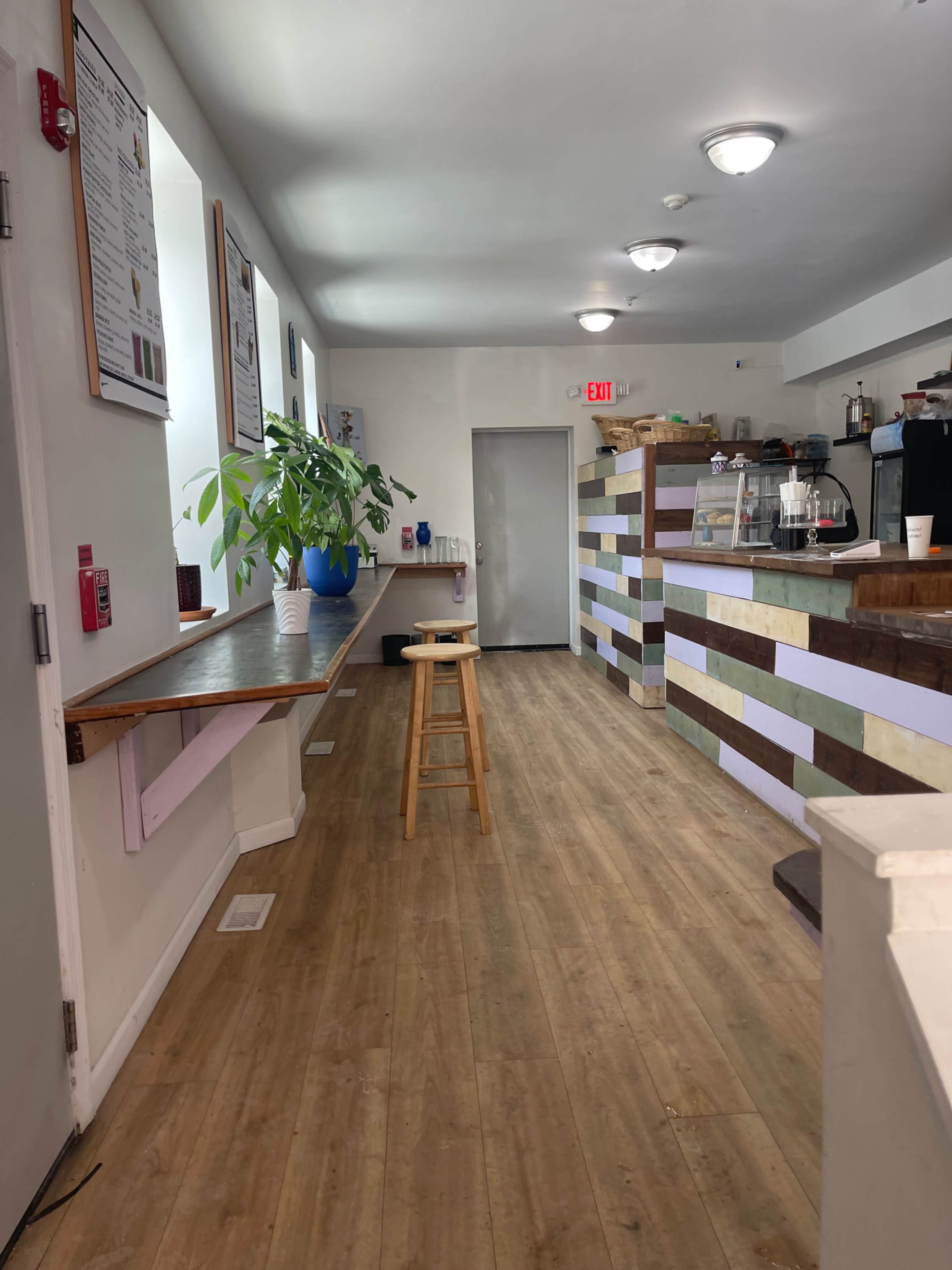The image shows a brightly lit café interior featuring a long table along the left side, a stool, and a colorful service counter at the far end.