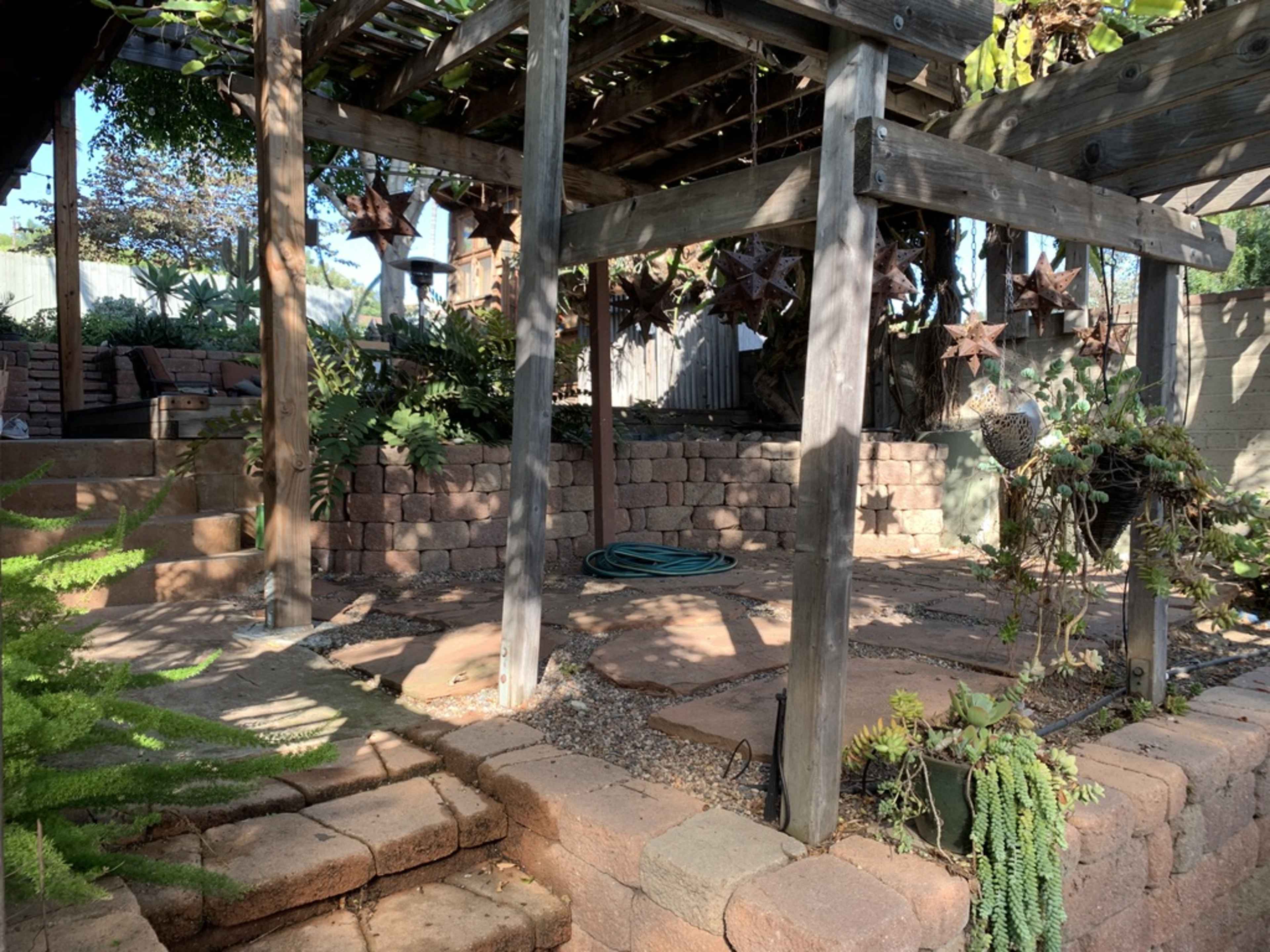 The image shows a landscaped garden area with stone pathways, wooden structures, and potted plants.