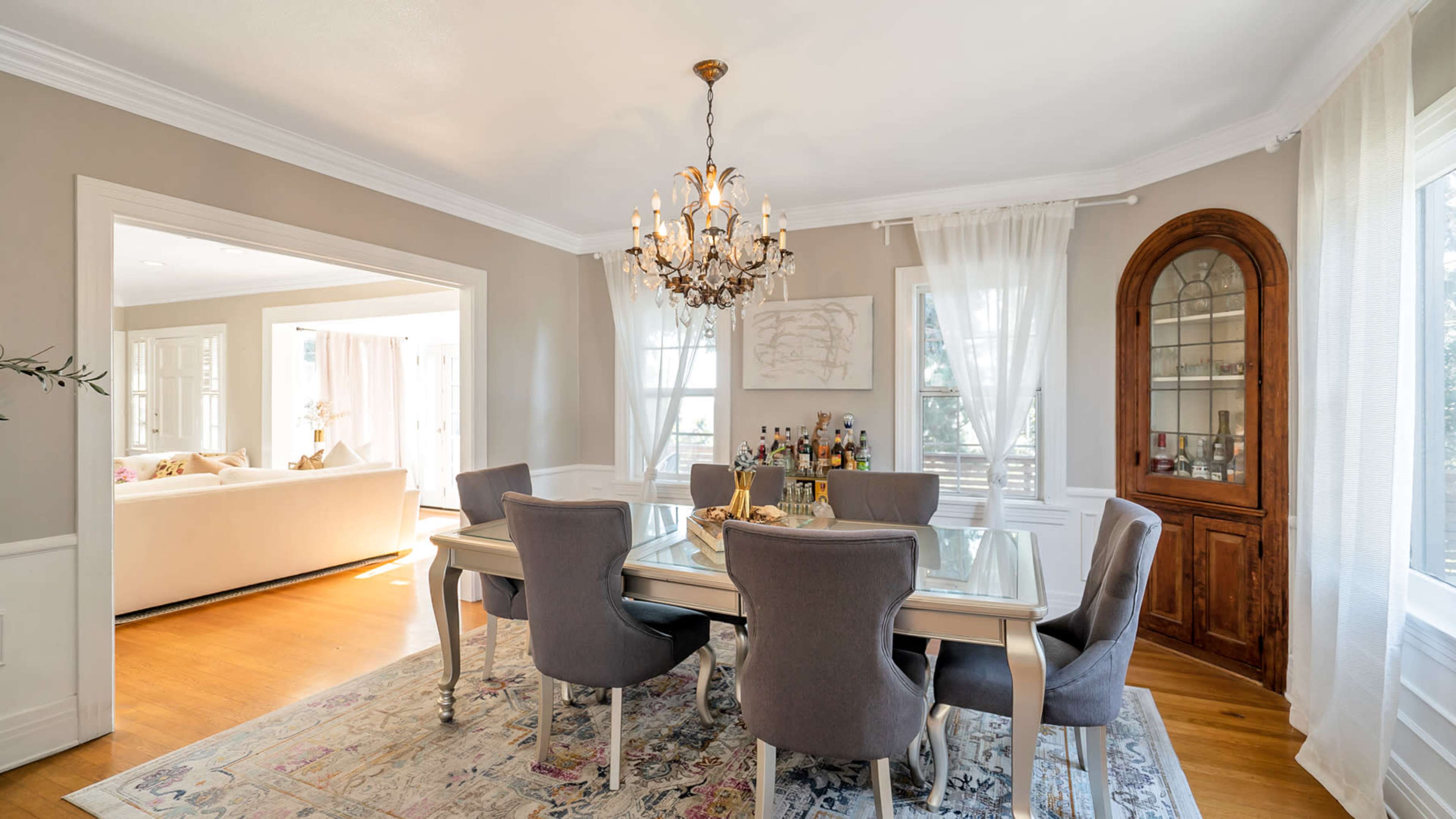 The dining room features a glass-topped table surrounded by gray upholstered chairs, with a chandelier overhead and a china cabinet against the wall.
