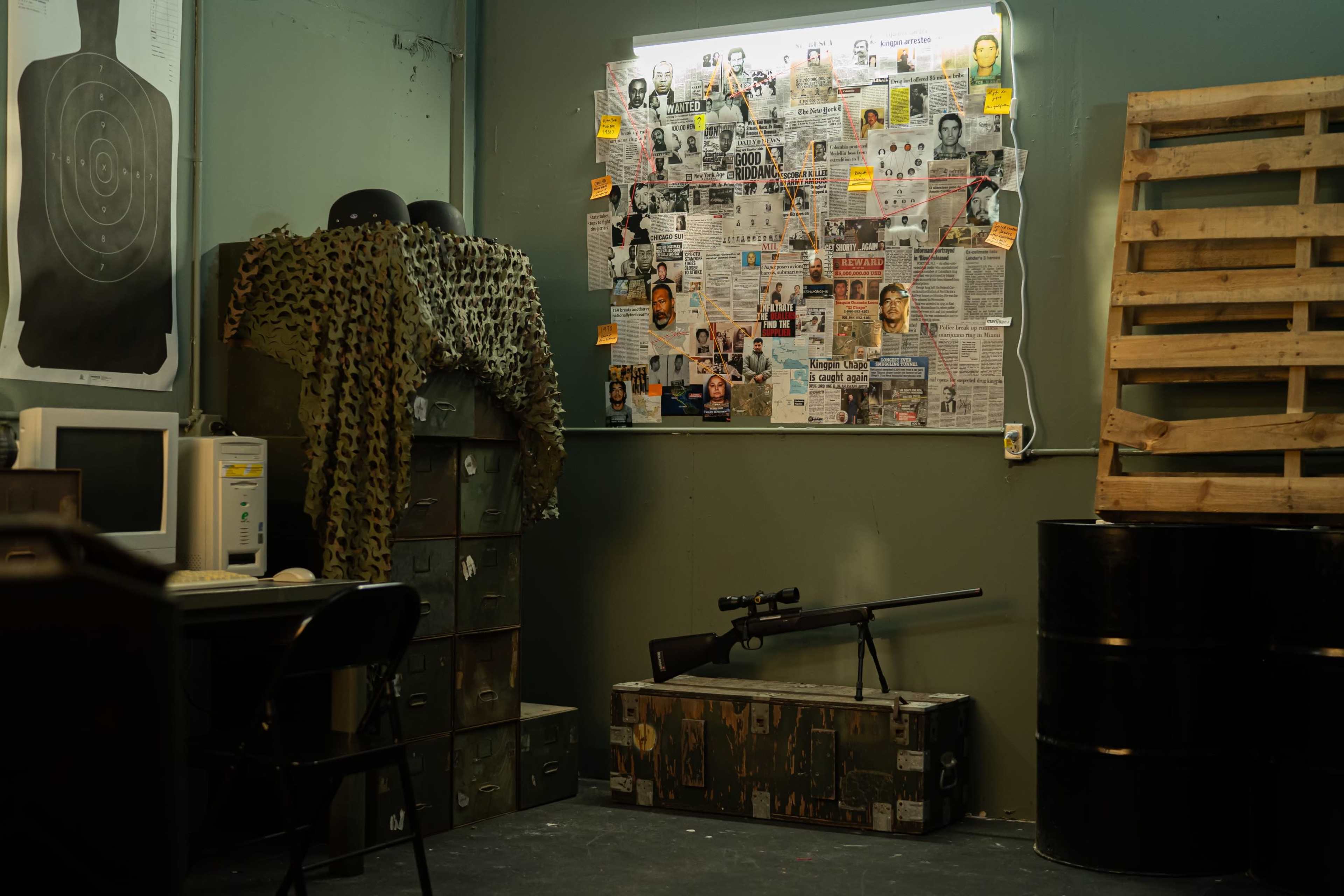A workspace featuring a desk with an old computer, a camouflage net draped over a stack of crates, and a wall covered with newspaper clippings and photographs, alongside a sniper rifle resting on a wooden crate.