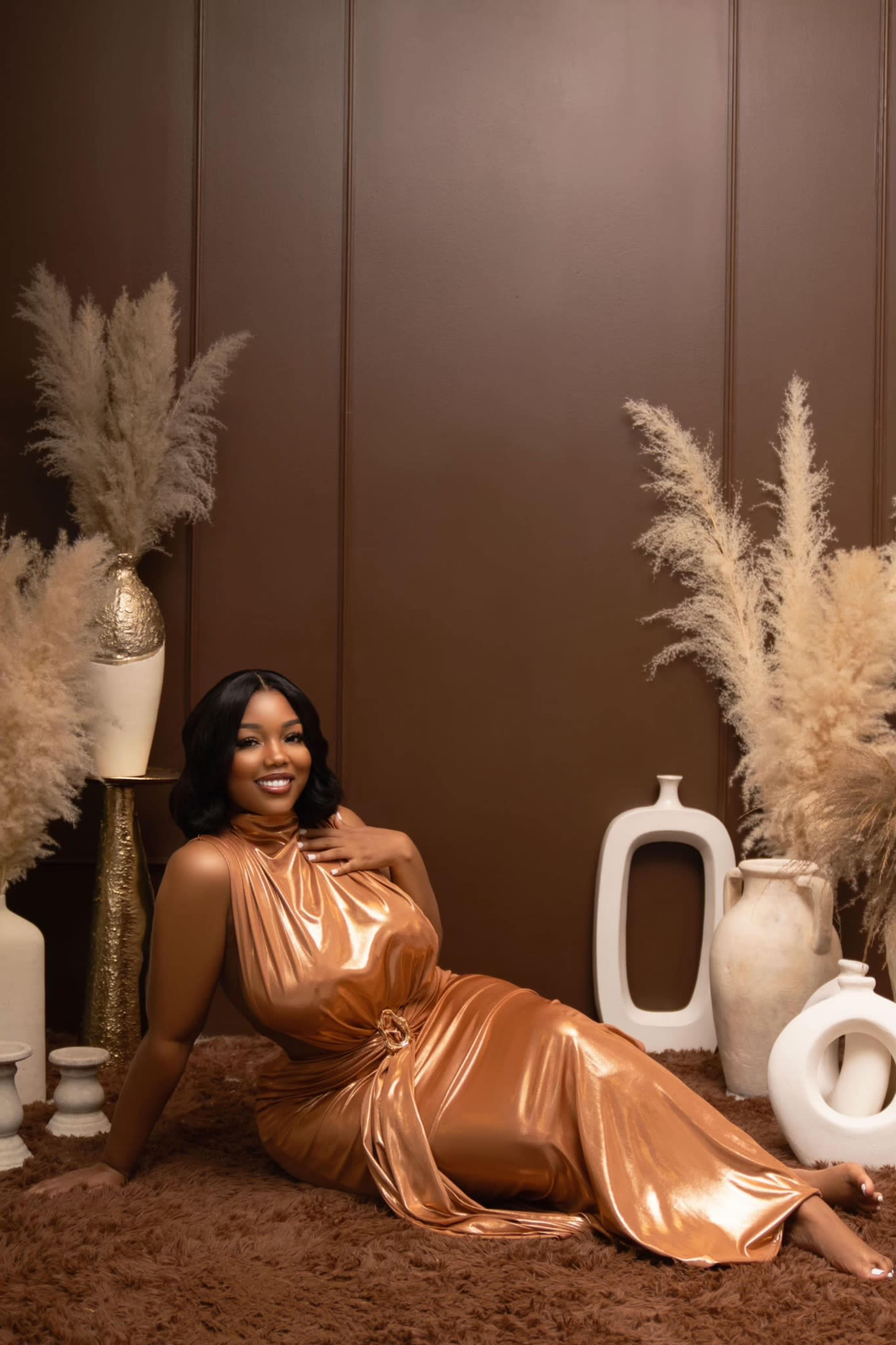 A person in a metallic copper dress is reclining on a brown rug surrounded by decorative vases and dried plants against a brown backdrop.