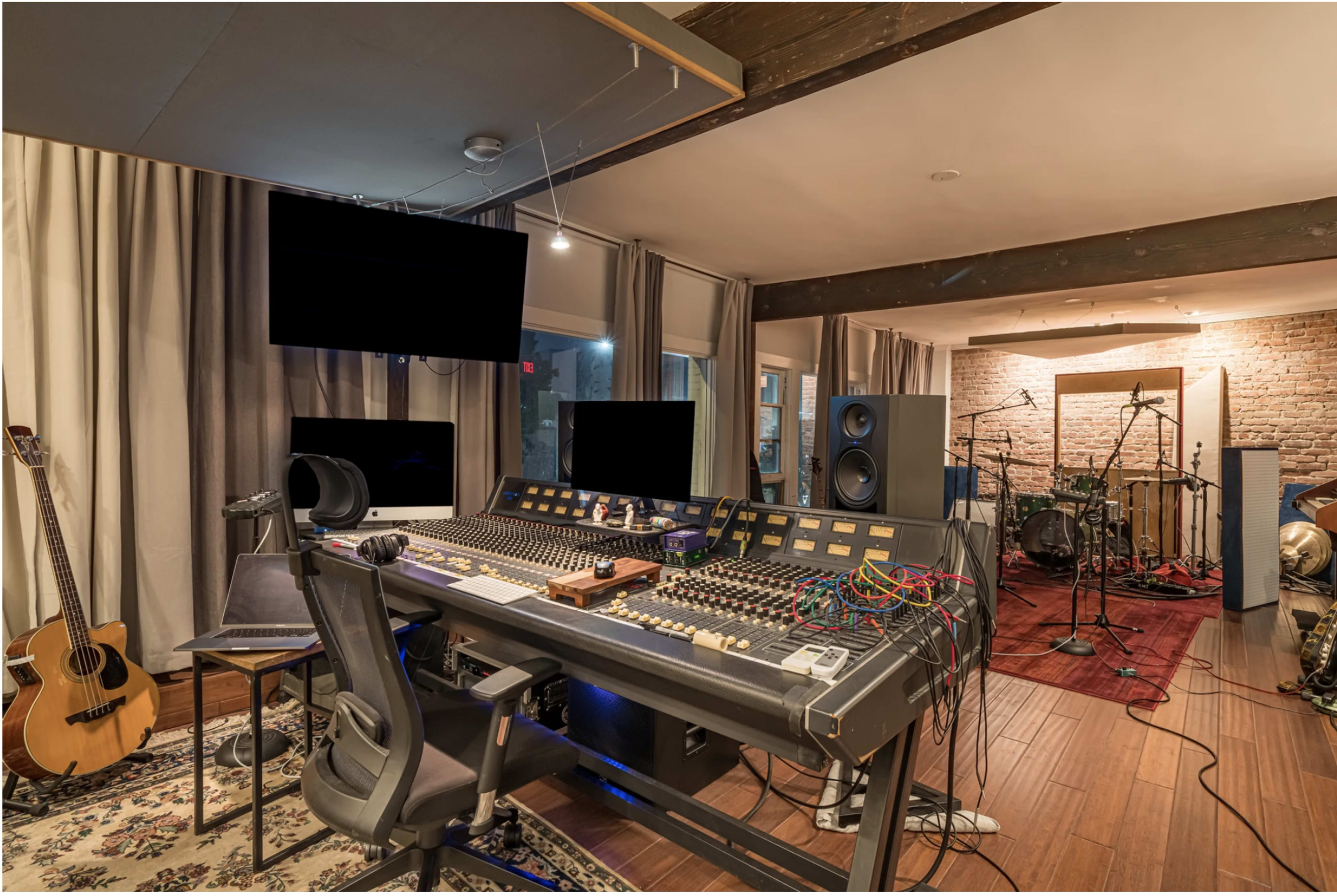 The image shows a recording studio with a large mixing console, several monitors, and various musical instruments including an acoustic guitar and drum setup.