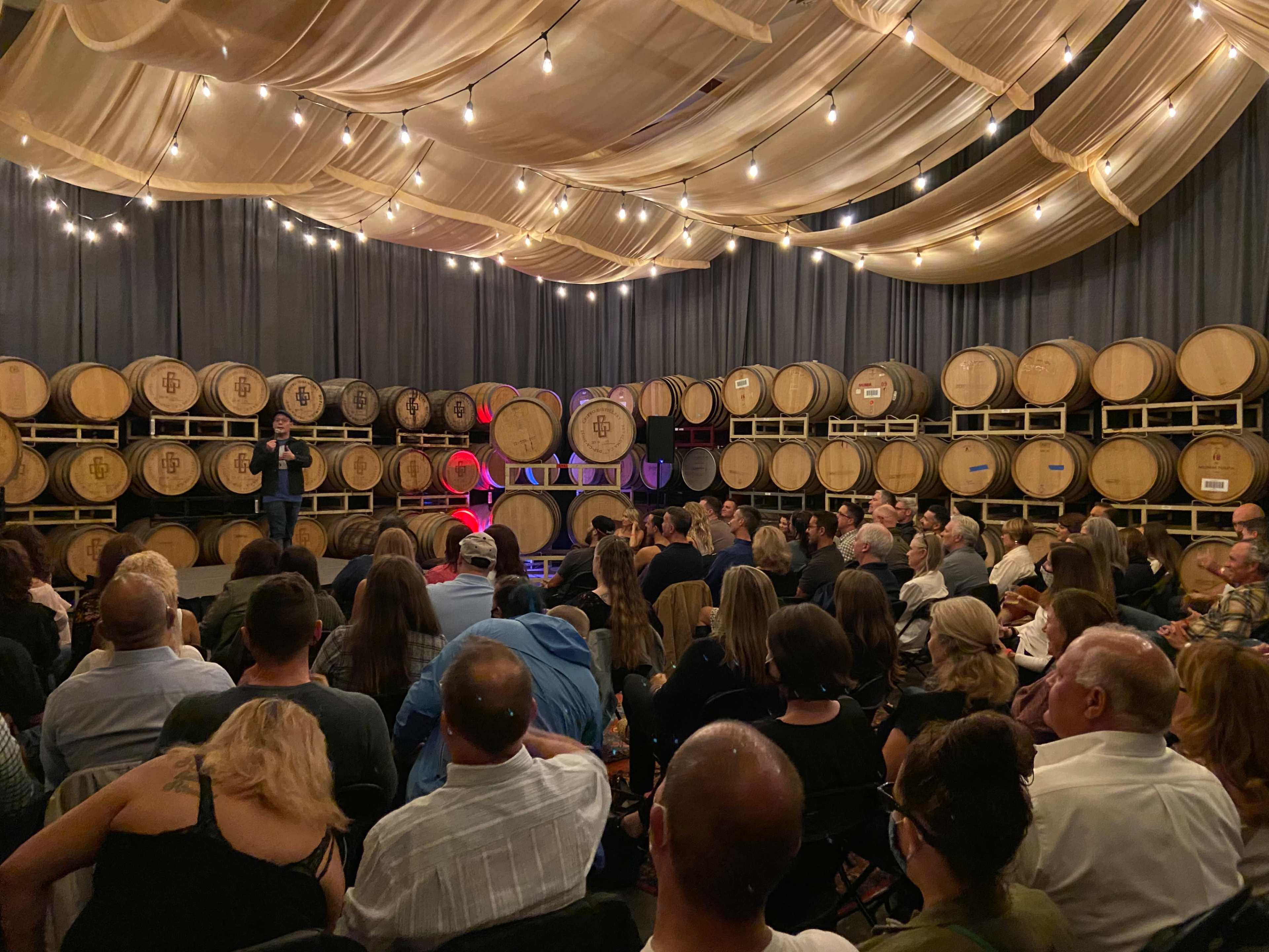 A speaker stands on a stage in a barrel room filled with seated audience members, illuminated by soft string lights overhead.