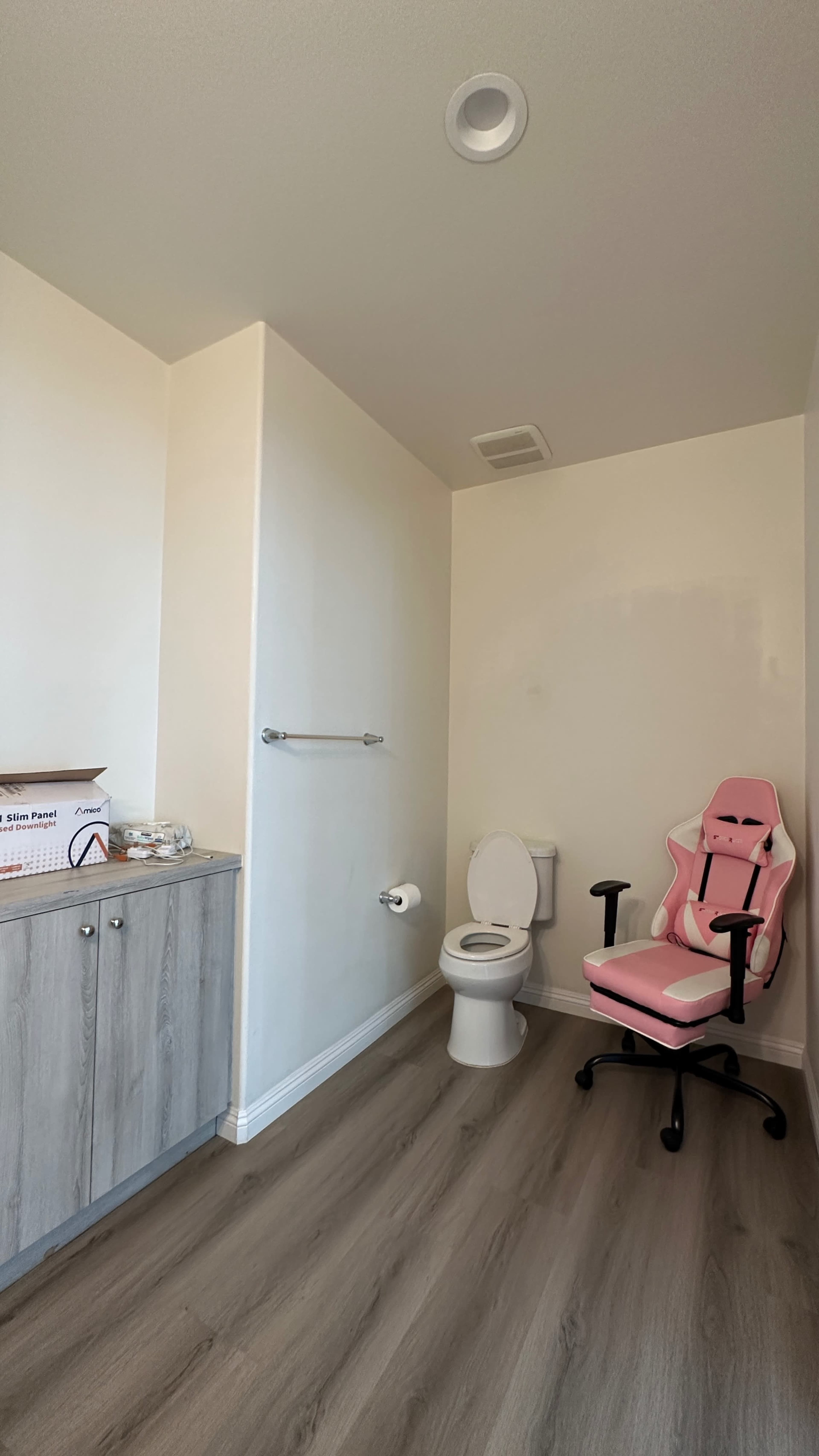 The room features a toilet, a pink gaming chair, and a cabinet against one wall with a towel rack nearby.