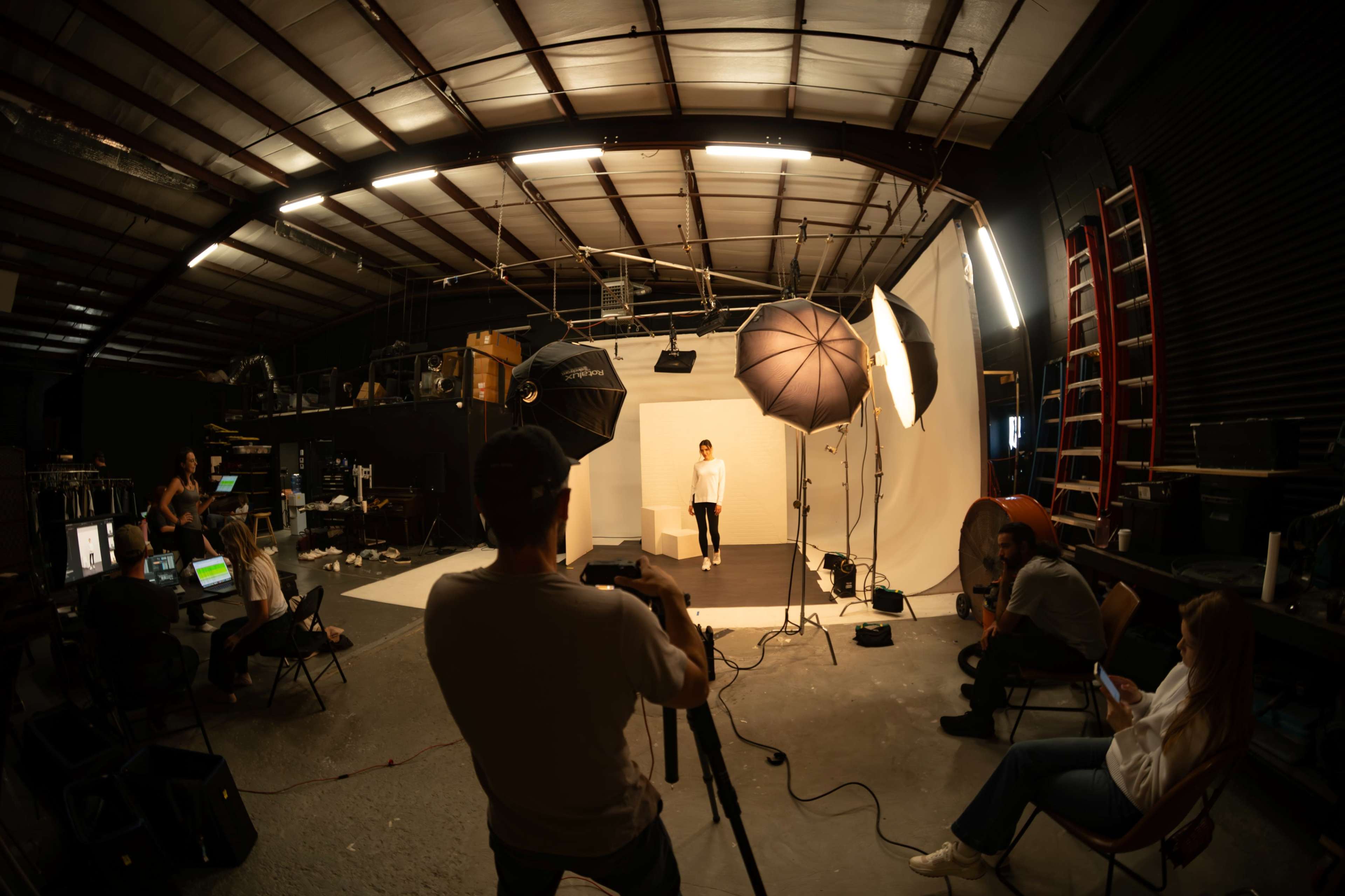 A photography studio is set up with multiple lights and a model posing against a white backdrop while a photographer captures the scene.