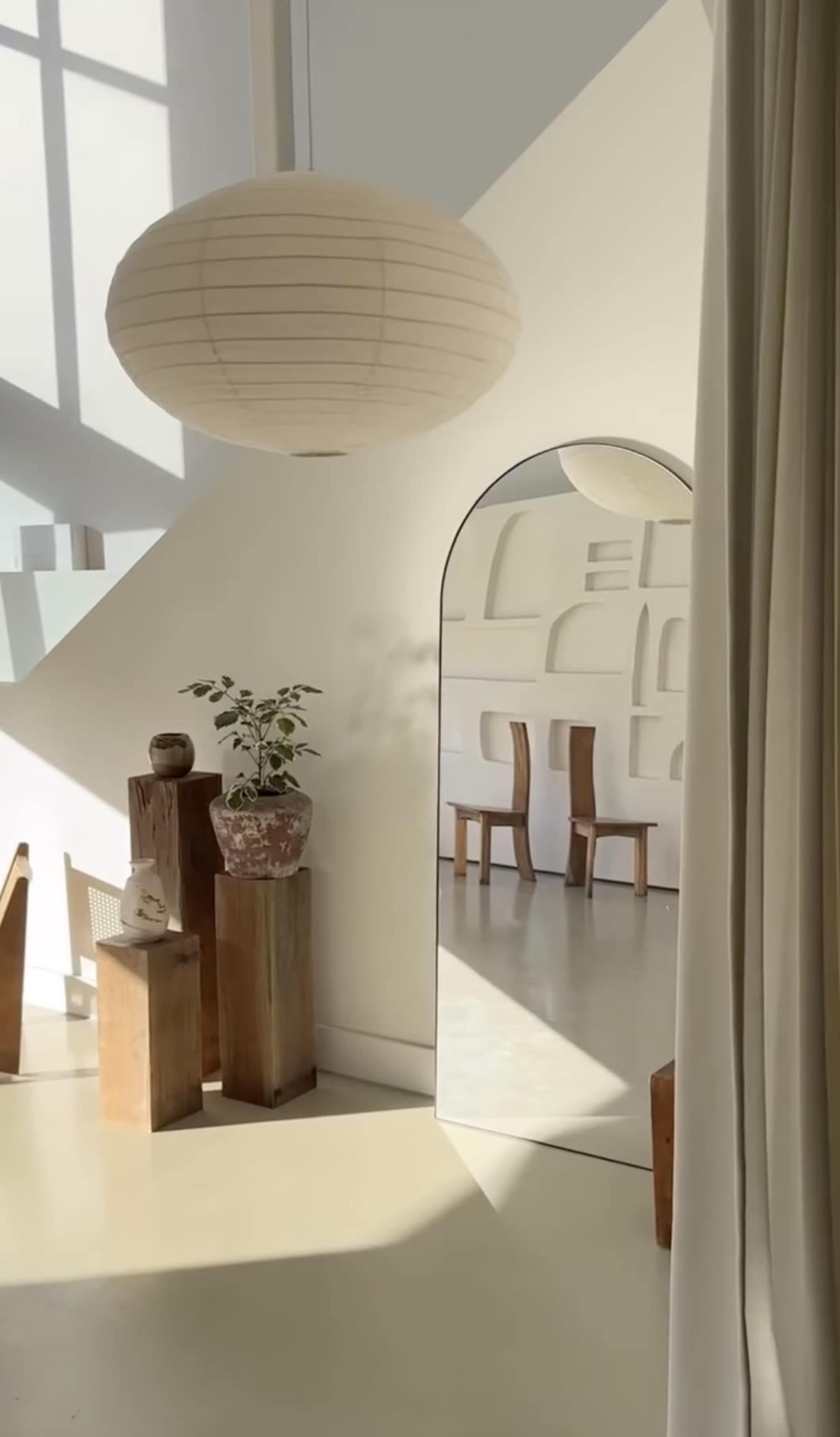 The image shows a bright interior space featuring a large round pendant light, wooden furniture, a decorative plant, and a mirror reflecting a minimalist wall design.